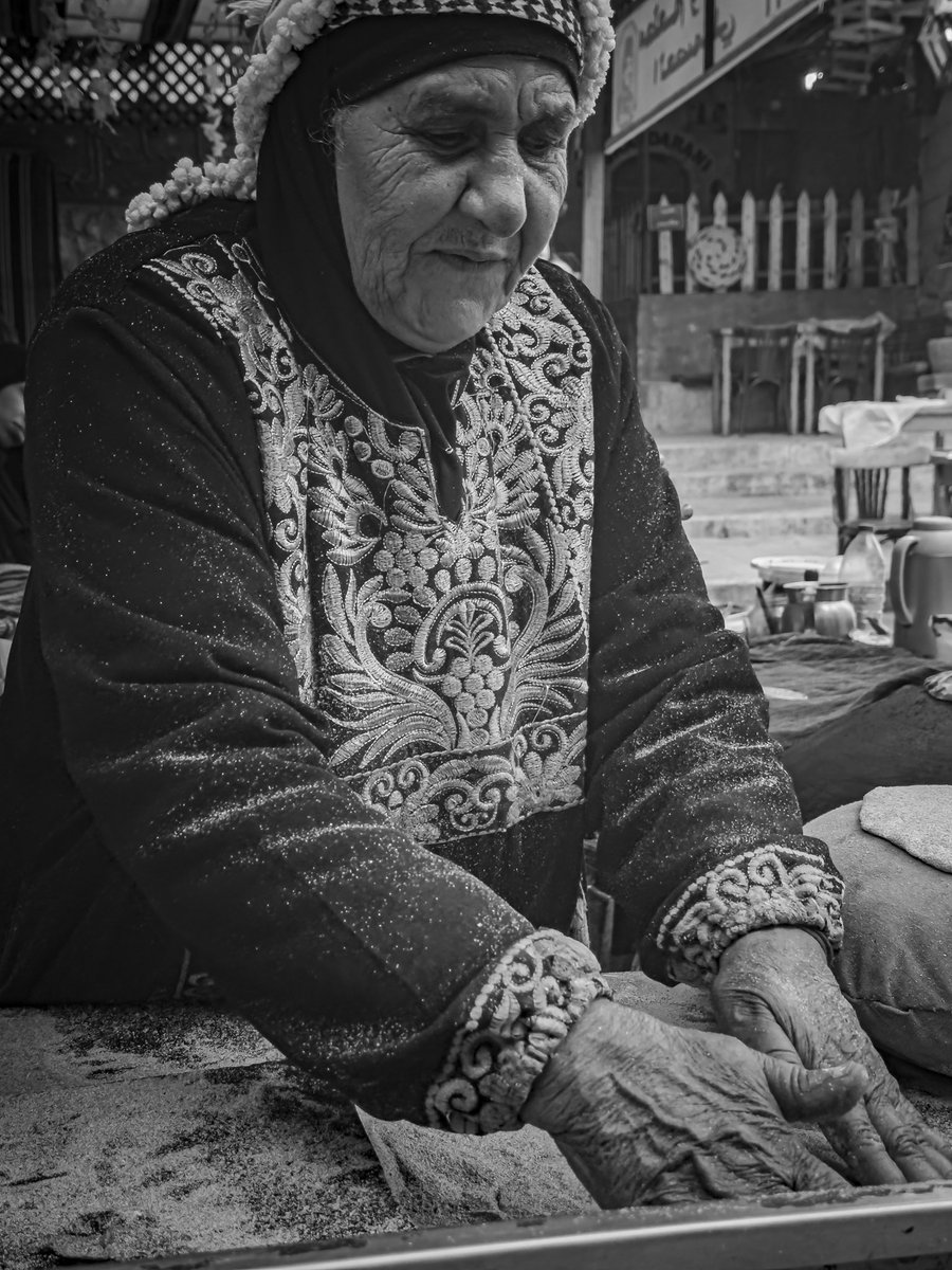 ndarwish's tweet photo. goodness - lived

in al-salt, between stone that has memorised generations, there are conversations that stay longer than photographs.

i met um ghazi.
she stood in the narrow street, freshly baking traditional sweets, not as a performance, but as continuation.
her presence did not ask for attention. it carried it.

passers by stopped. not only for the sweets made, but for the directness in her gaze, the kind that does not pretend. the kind that has seen enough to speak plainly.

i asked her one question.
what makes a good person these days?

she did not hesitate. “اخلاقه”. his manners. his character. the way that one conducts themselves when no one is measuring.
then she added what only someone seasoned by life would say. “اتقِ شر من أحسنت إليه”.
be mindful, even of the one you were good to.
not fear. awareness.
because goodness is not blindness. it is clarity with restraint.
it is knowing that kindness does not promise return, and choosing it anyway.

there are people who have witnessed enough to harden.
yet they refuse to.
that refusal is not softness.
it is discipline of the soul.
character that does not bend,
simply because the world does.

___________________________
#streetphotography by nabil darwish, copyright 2026 all rights reserved.
location: #alsalt, #jordan
___________________________

#storytelling, #visualstorytelling, #lensculture, #natgeocreative, #natgeotravel, #photography, #people, #natgeo, #worldpressphoto, #magnumphotos, #portrait, #opensociety, #vii, #viiphoto, #documentaryphotography, #storytelling, #thephotosociety, #art, #portraitphotography, #yourshotphotographer, #noorimages, #visualart, #life, #blackandwhite, #streetportrait