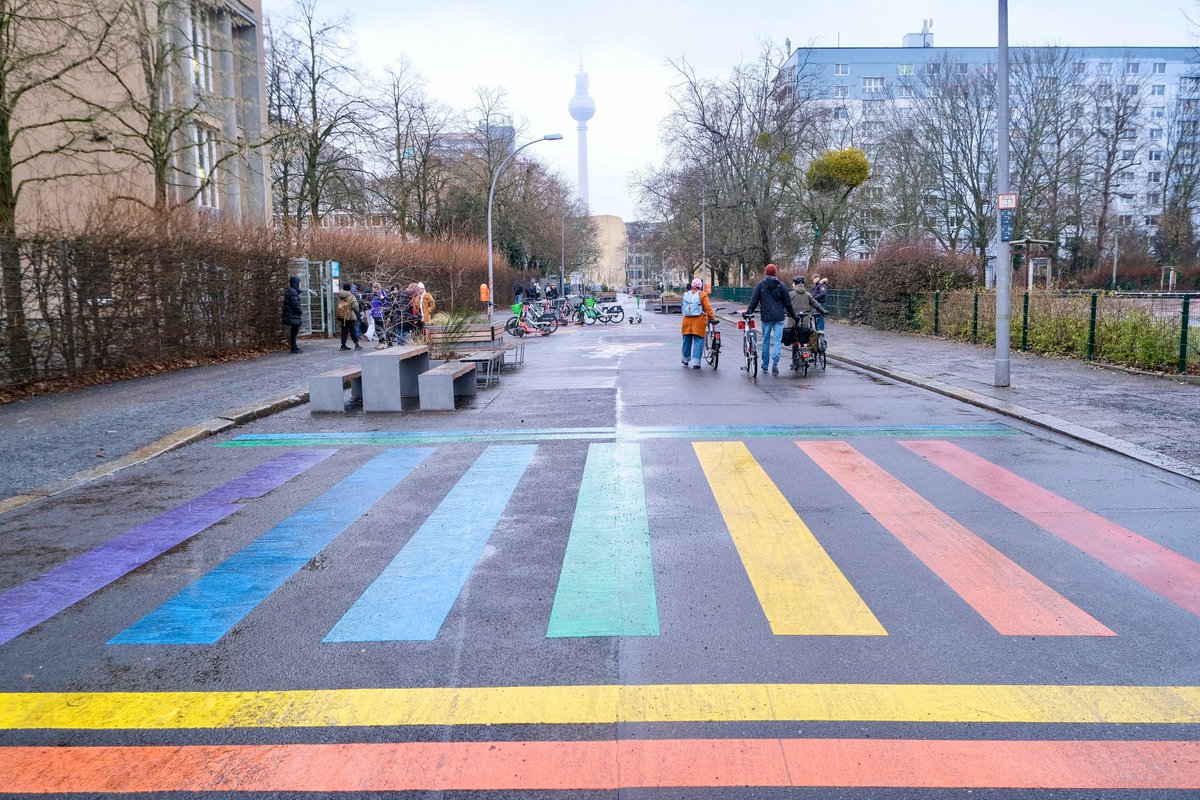 People in casual winter attire walk, cycle, stand, and sit in a Berlin Kiezblock or "neighbourhood block". The traffic filtered street or intersection contains bollbards, planter boxes, parklets, playground equipment, painted murals, and park benches, and is lined with trees and apartment buildings.