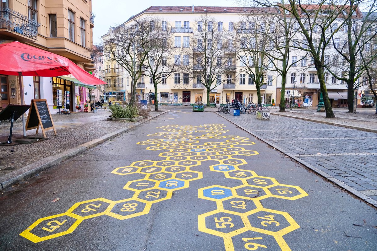People in casual winter attire walk, cycle, stand, and sit in a Berlin Kiezblock or "neighbourhood block". The traffic filtered street or intersection contains bollbards, planter boxes, parklets, playground equipment, painted murals, and park benches, and is lined with trees and apartment buildings.