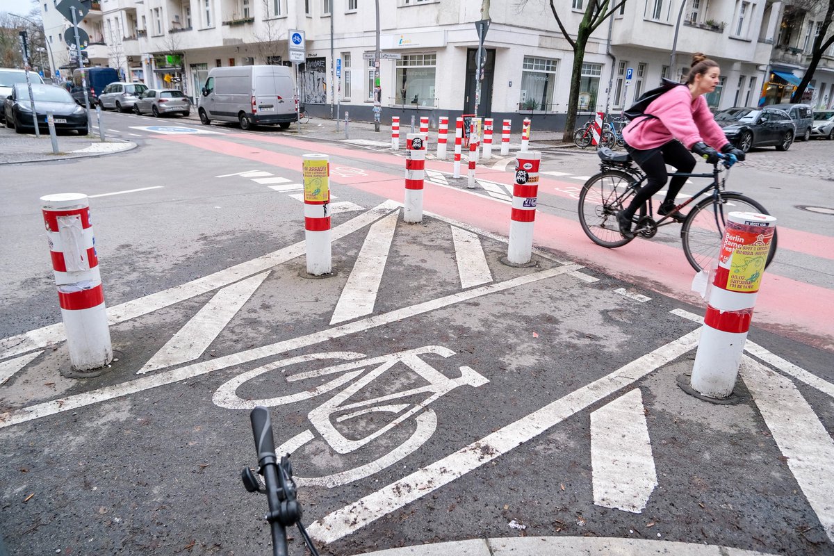 People in casual winter attire walk, cycle, stand, and sit in a Berlin Kiezblock or "neighbourhood block". The traffic filtered street or intersection contains bollbards, planter boxes, parklets, playground equipment, painted murals, and park benches, and is lined with trees and apartment buildings.