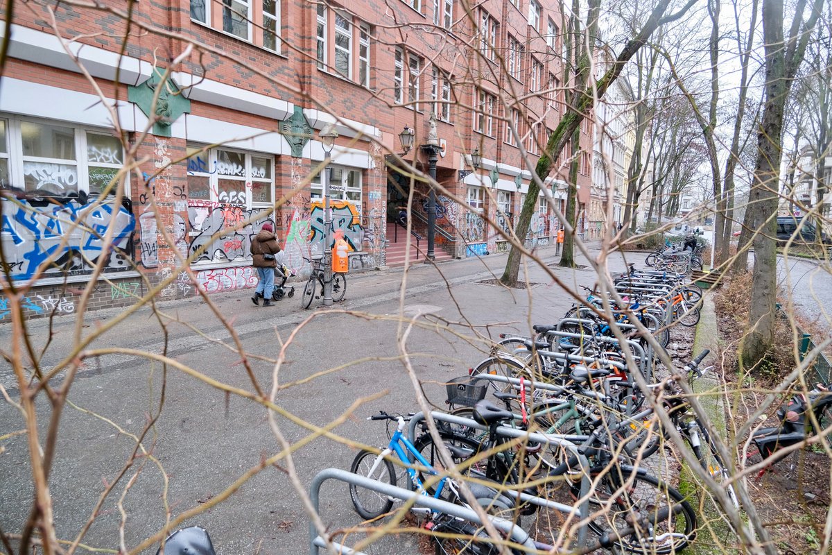 People in casual winter attire walk, cycle, stand, and sit in a Berlin Kiezblock or "neighbourhood block". The traffic filtered street or intersection contains bollbards, planter boxes, parklets, playground equipment, painted murals, and park benches, and is lined with trees and apartment buildings.