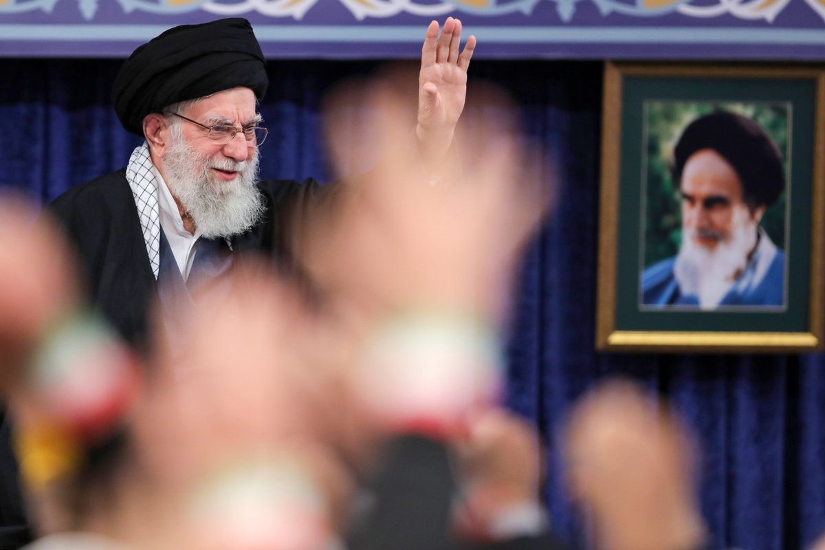 Ayatollah Ali Khamenei smiling and waving with right hand raised to a crowd in a meeting hall, blurred audience hands in foreground, framed portrait of a bearded man on blue curtained wall behind.