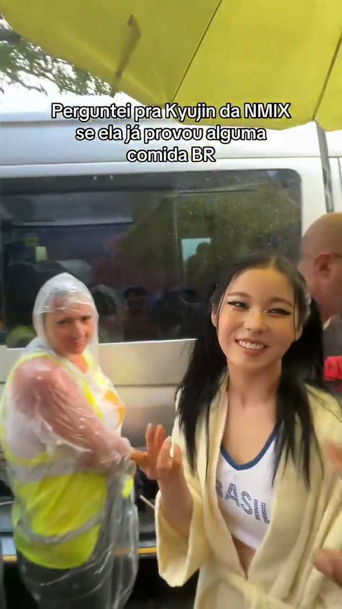 Imagen con texto en portugués 'Perguntei pra Kyujin da NMIXX se ela já provou alguma comida BR' mostrando a la idol Kyujin sonriendo bajo un paraguas amarillo.