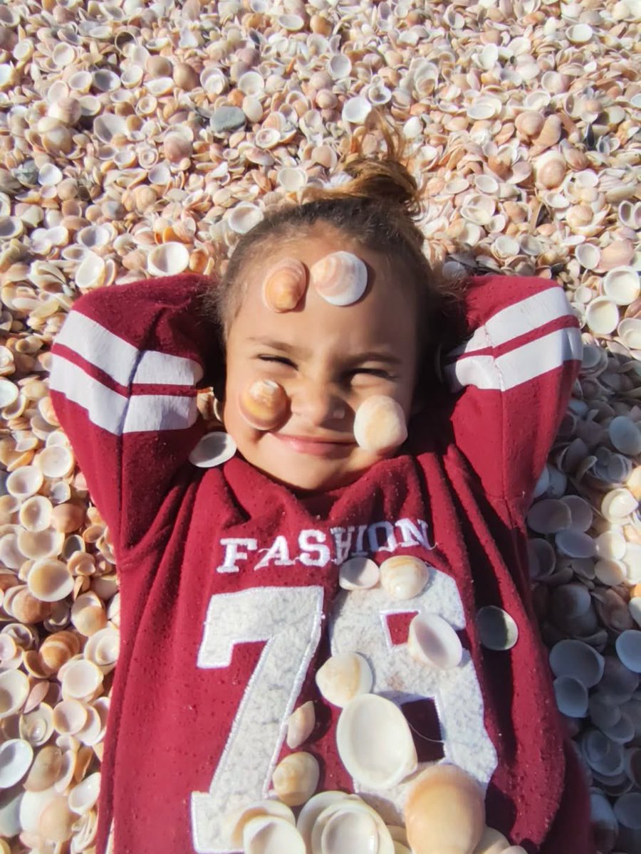 Gazze sahilinde, deniz kabukları içinde gülümseyen Gazzeli bir kız çocuğu. ✨ 

4 yaşındayım, hâlâ hayal ediyorum, oyunlar oynuyorum.
Bu deniz, bu toprak, bu hava bizim diyor. ❤️‍🩹