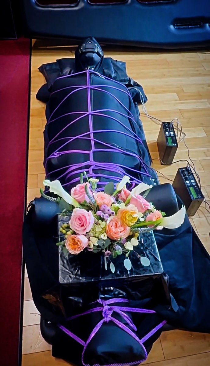 Slave in a Darlexx Sleepsack with crotch length bespoke Fernando Berlin Boots, leather jumpsuit made in India, Mr S Leather flat panel hood. ErosTek 312 boxes provide Estim to his nether regions as well as nipples. The floral arrangement he brings each visit is on display.