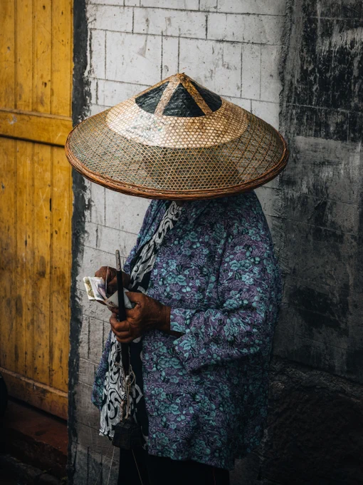 An individual in traditional Asian attire stands against a textured wall. They wear a conical hat obscuring their face and a floral-patterned top. Their hands hold a fan of paper money. A yellow door is visible in the background.
