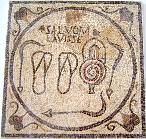 This 3rd century Roman floor mosaic at an entrance to public bathhouse depicts a pair of bathing sandals, an oil flask and three curved “strigils” used for scraping off sweat, dirt and oil. The inscription declares that bathing is good for you (Sabratha Museum, Libya)