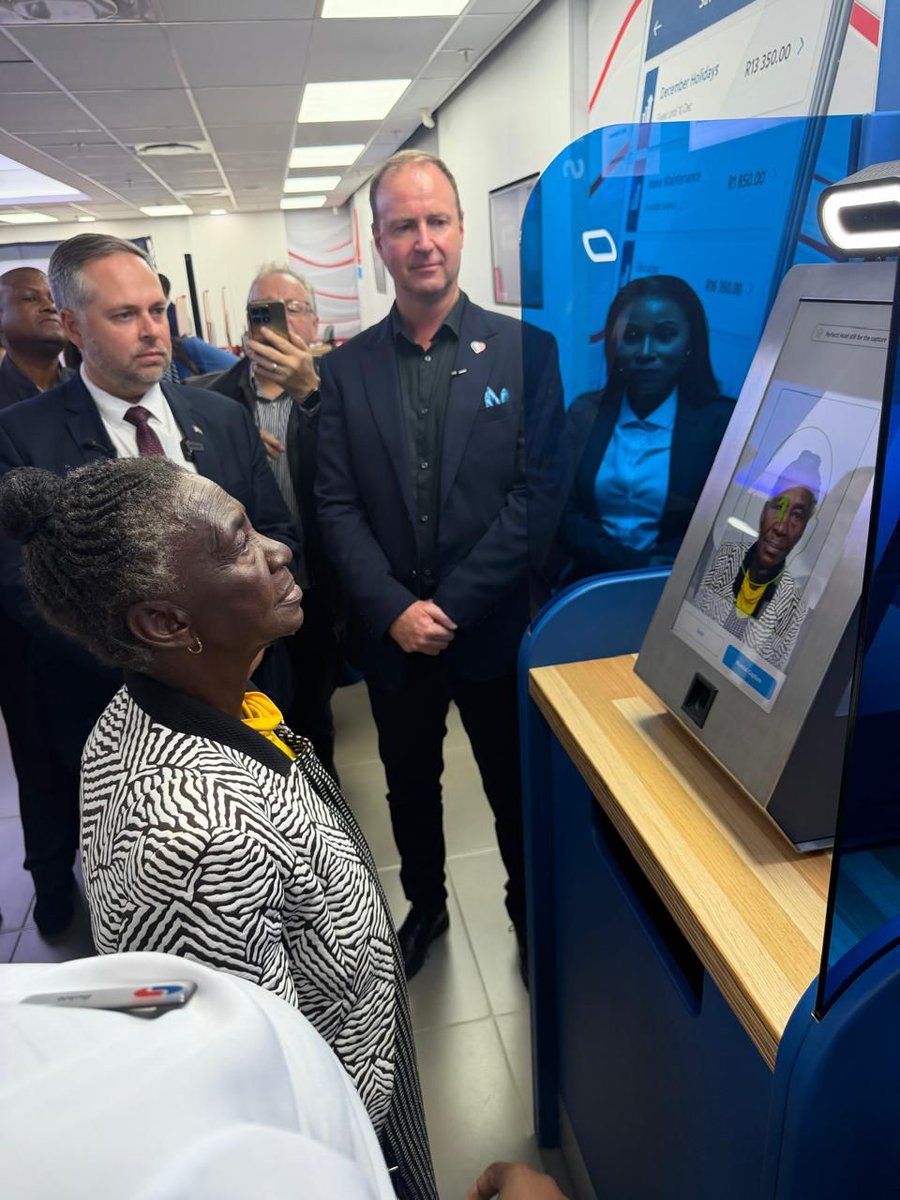 Elderly woman in patterned jacket at blue Smart ID kiosk her photo on screen men in suits watch