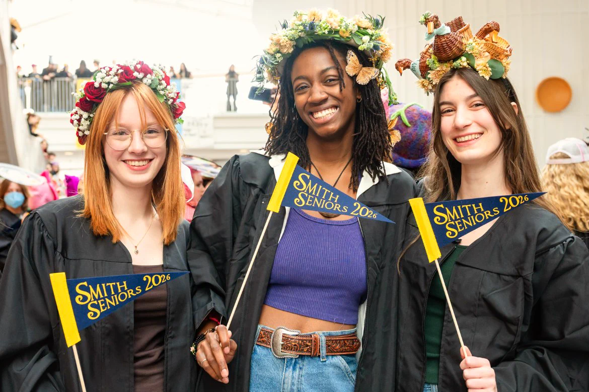 A group of three graduates in black gowns, each holding a small flag that says "Smith Seniors 2026." They are standing indoors, smiling, and wearing creative hats.