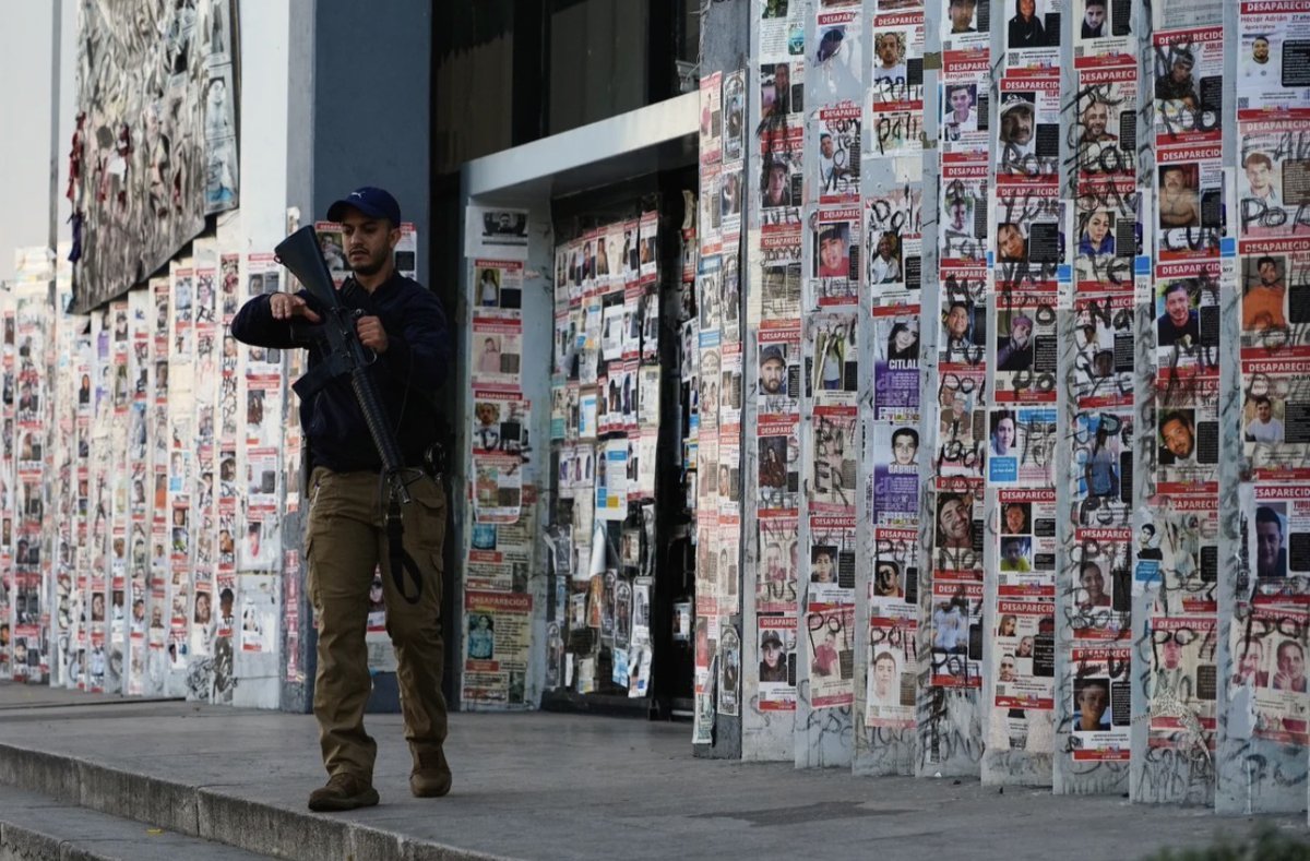 The image shows a man holding a rifle walking past a wall covered with numerous posters, each depicting photographs of people and text.