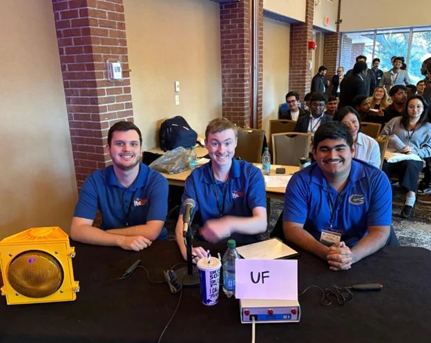 Kyle Groome, Michael Panarisi, and Hugo Resendez are sitting at a table with microphones and a sign labeled "UF". They are wearing matching blue shirts with logos. An audience is seated behind them, visible in the background.