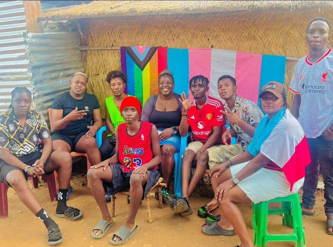 QueerCliff's tweet photo. 🌈In the heart of Gorom Refugee Settlement (first photo), where safety is never guaranteed and every day demands courage, we still find ways to gather, to breathe, to be fully ourselves.
In the second photo, our community, back then in Kakuma refugee camp, stood strong on dusty grounds of the Turkana desert, rainbow colors shining in clothing and spirit, even as the police came in trucks to brutally crash the peaceful protest against homophobia that we had organized that day. This isn't just a photo. It's a statement that amid uncertainty, threats, and the constant weight of displacement, we refuse to hide or breakdown. We stand together, visible, unapologetic, saying "No" to homophobia with every outfit, every linked arm, every raised voice. "SAY NO TO HOMOPHOBIA" isn't just printed on a shirt, it is lived here, every single day.
The first photo brings it home: friends and chosen family squeezed onto stools under a thatched roof, a vibrant pride flag backdrop framing our smiles, peace signs, and laughter. We share space, stories, and hope. In a place where basic needs are stretched thin and resettlement feels distant, these moments remind us why we keep going: because community is our strongest shelter.
From Kakuma's dangers to Gorom's challenges, we have crossed borders, survived violence, and rebuilt again and again. Homophobia follows, aid shortages bite harder, and the world often looks away... but look at us. We are still here. Still laughing. Still loving. Still raising our flags against the odds.
This is queer resilience in its rawest form. This is chosen family refusing to be broken. This is proof that joy can bloom even in the harshest soil.

We exist. We resist. We shine. 🌈🫂💪
#QueerRefugees #GoromCamp #LGBTQRefugees #PrideInDisplacement #RainbowFamily #FromKakumaToGorom #QueerJoyIsResistance #EastAfricaQueer #ResilientAndRainbow #SayNoToHomophobia