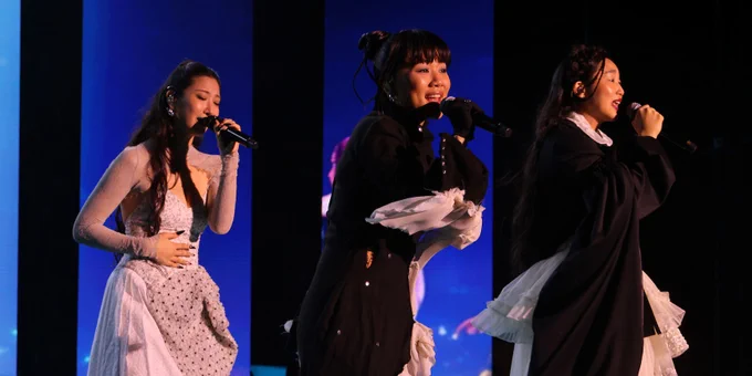 A wide shot of a vocal group HUNTR/X performing on stage. Rei Ami  ears a long black coat with a white ruffled train and black boots. To her left, EJAE wears a white lace dress with sheer sleeves and Audrey Nuna wears a voluminous black and white layered ensemble with white platform shoes. All three are holding microphones and singing under blue stage lights.