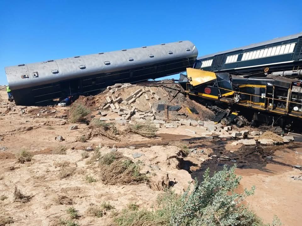 Silver passenger car overturned on dirt mound amid debris in desert under blue sky