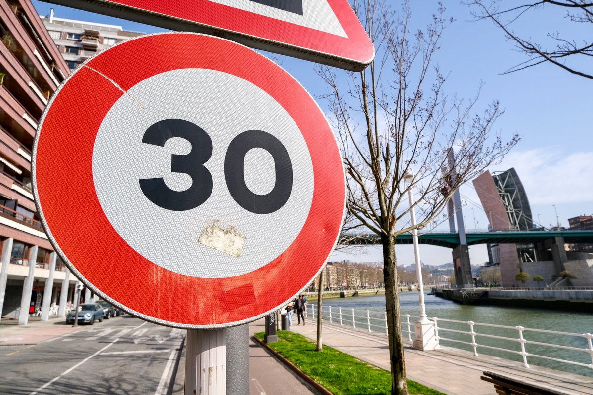 People of all ages and abilities walk, cycle, drive, and ride public transportation along the low-traffic streets of Bilbao, which are lined with buildings, trees, bike infrastructure, and prominent 30 km/h speed limit signage and road markings.