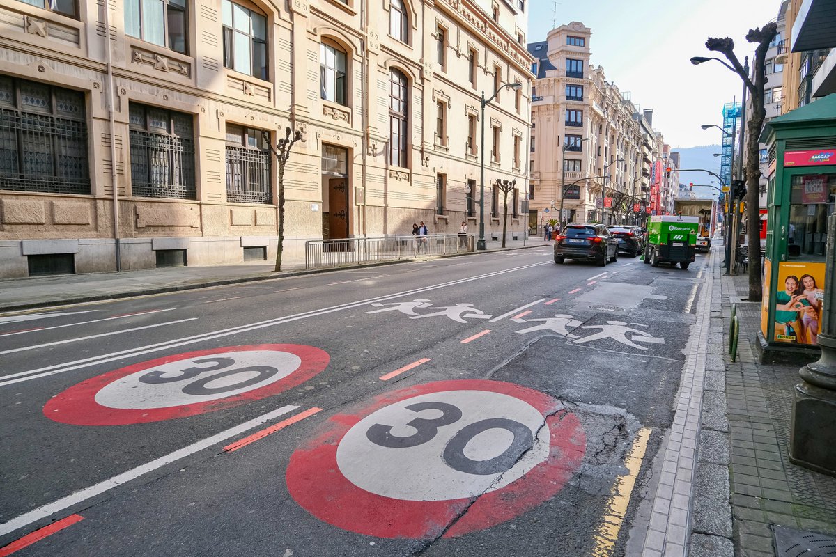 People of all ages and abilities walk, cycle, drive, and ride public transportation along the low-traffic streets of Bilbao, which are lined with buildings, trees, bike infrastructure, and prominent 30 km/h speed limit signage and road markings.