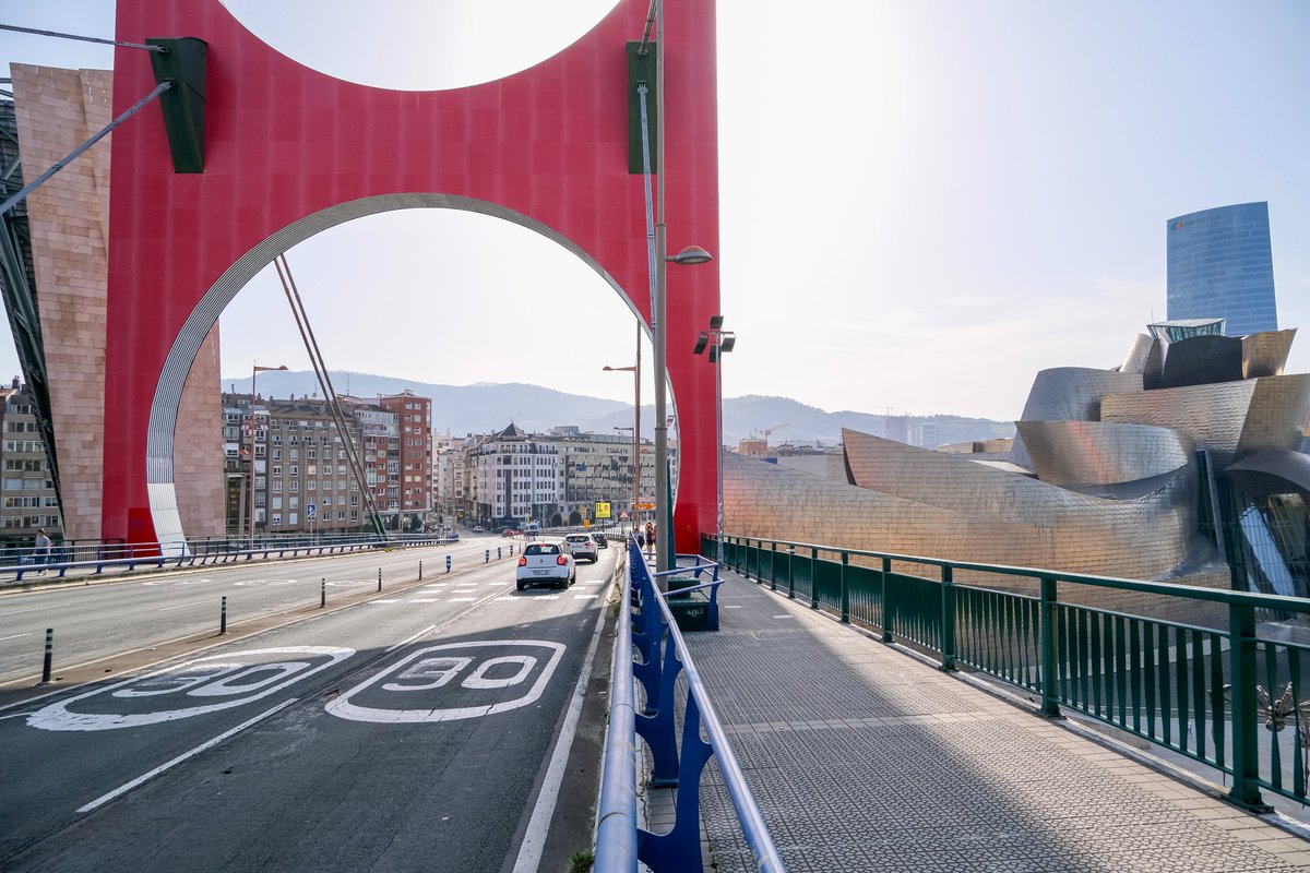 People of all ages and abilities walk, cycle, drive, and ride public transportation along the low-traffic streets of Bilbao, which are lined with buildings, trees, bike infrastructure, and prominent 30 km/h speed limit signage and road markings.