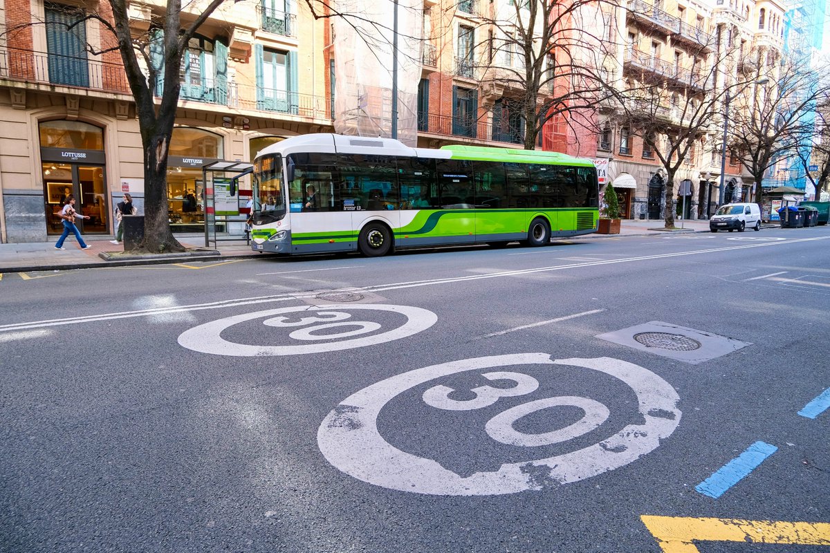 People of all ages and abilities walk, cycle, drive, and ride public transportation along the low-traffic streets of Bilbao, which are lined with buildings, trees, bike infrastructure, and prominent 30 km/h speed limit signage and road markings.