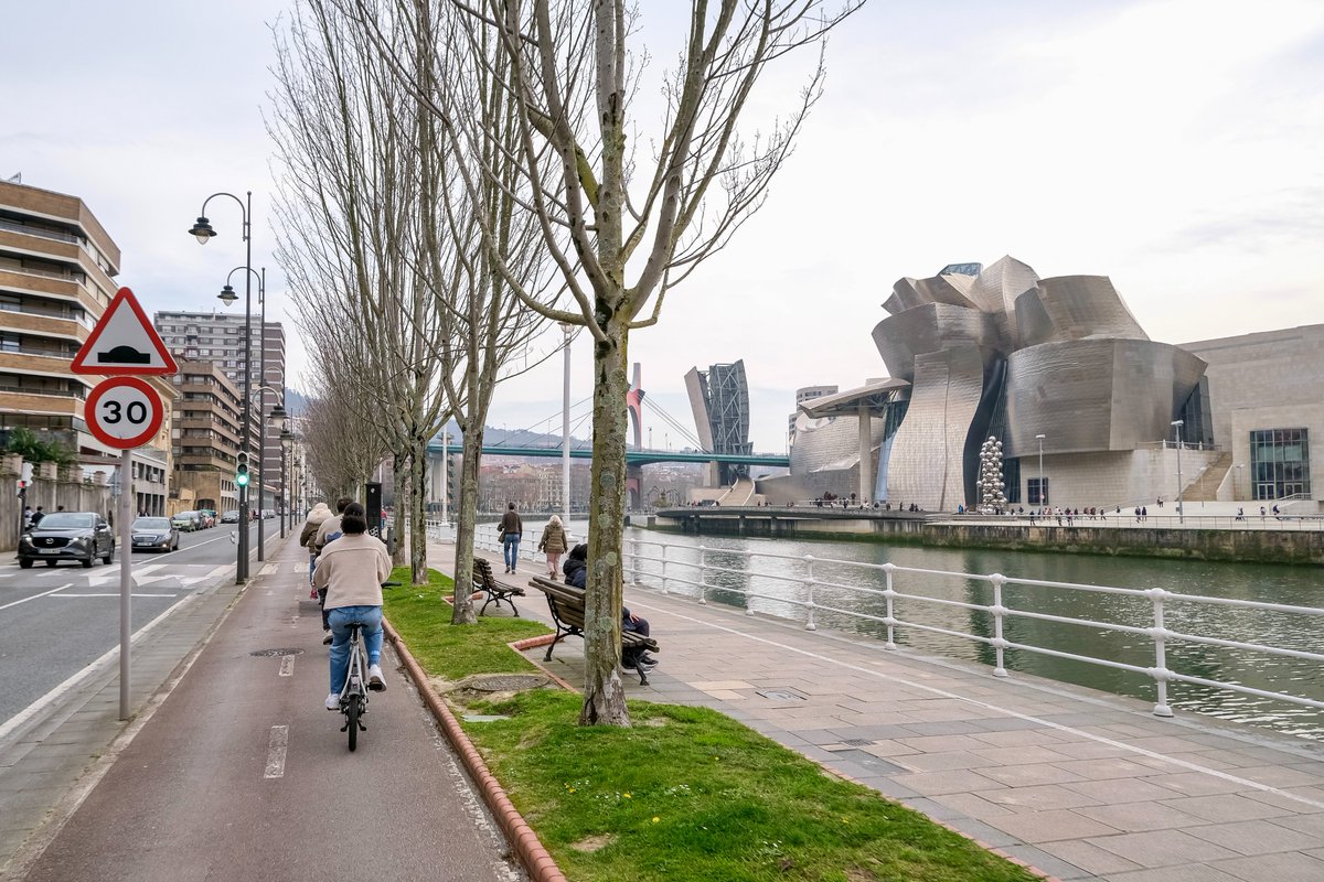 People of all ages and abilities walk, cycle, drive, and ride public transportation along the low-traffic streets of Bilbao, which are lined with buildings, trees, bike infrastructure, and prominent 30 km/h speed limit signage and road markings.