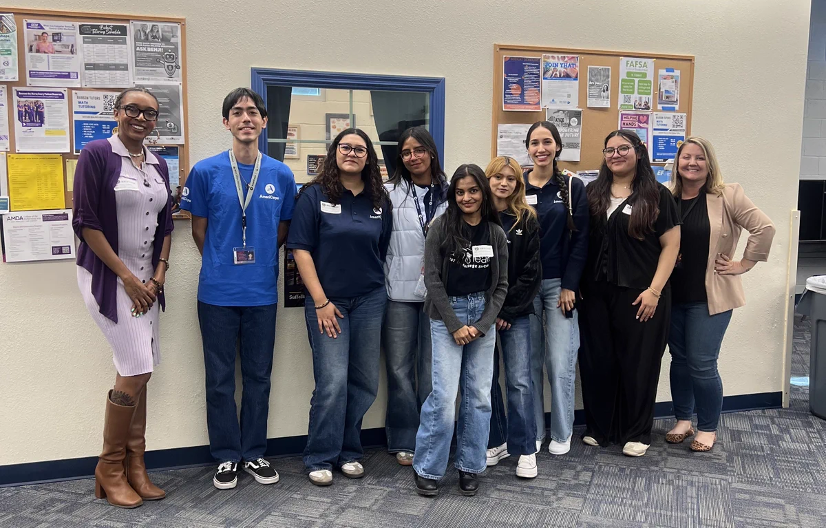 A group of nine diverse students and staff pose in a brightly lit office or classroom. Several wear blue AmeriCorps shirts. They stand in front of a blue-framed window and two bulletin boards covered in academic posters, including one for FAFSA. The group is dressed in casual business and student attire, ranging from jeans and t-shirts to a lavender dress with tall boots.