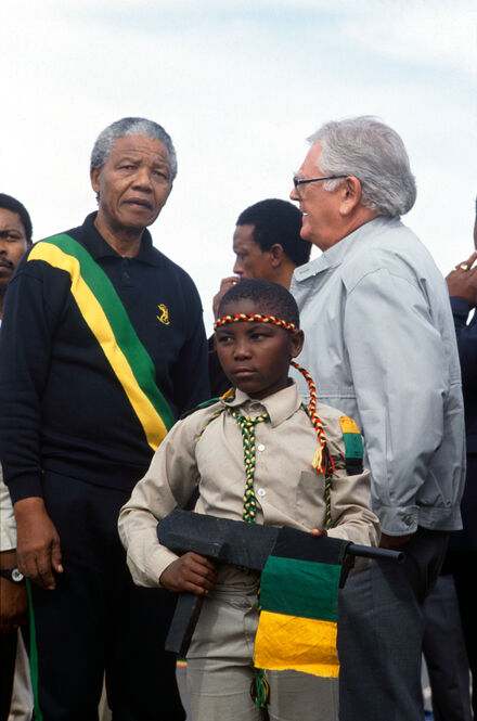 c 1990s: Nelson Mandela in ANC colours and Joe Slovo, leader of the South African Communist Party (SACP) at a rally. In front stands a young boy, dressed in ANC colours, holding a wooden rifle.  Photo Credit: Ken Oosterbroek