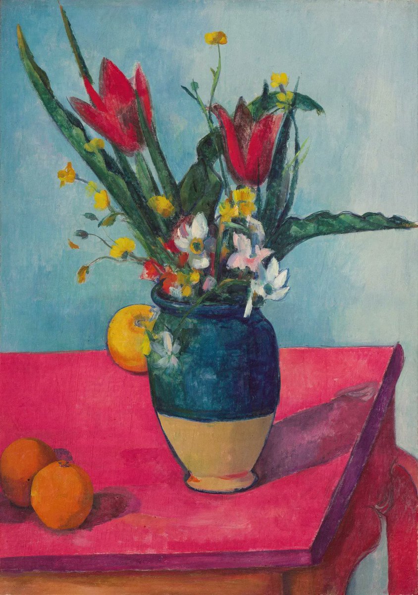 A bouquet of red flowers and yellow blooms in a blue vase sits on a vibrant pink tablecloth, accompanied by two oranges.