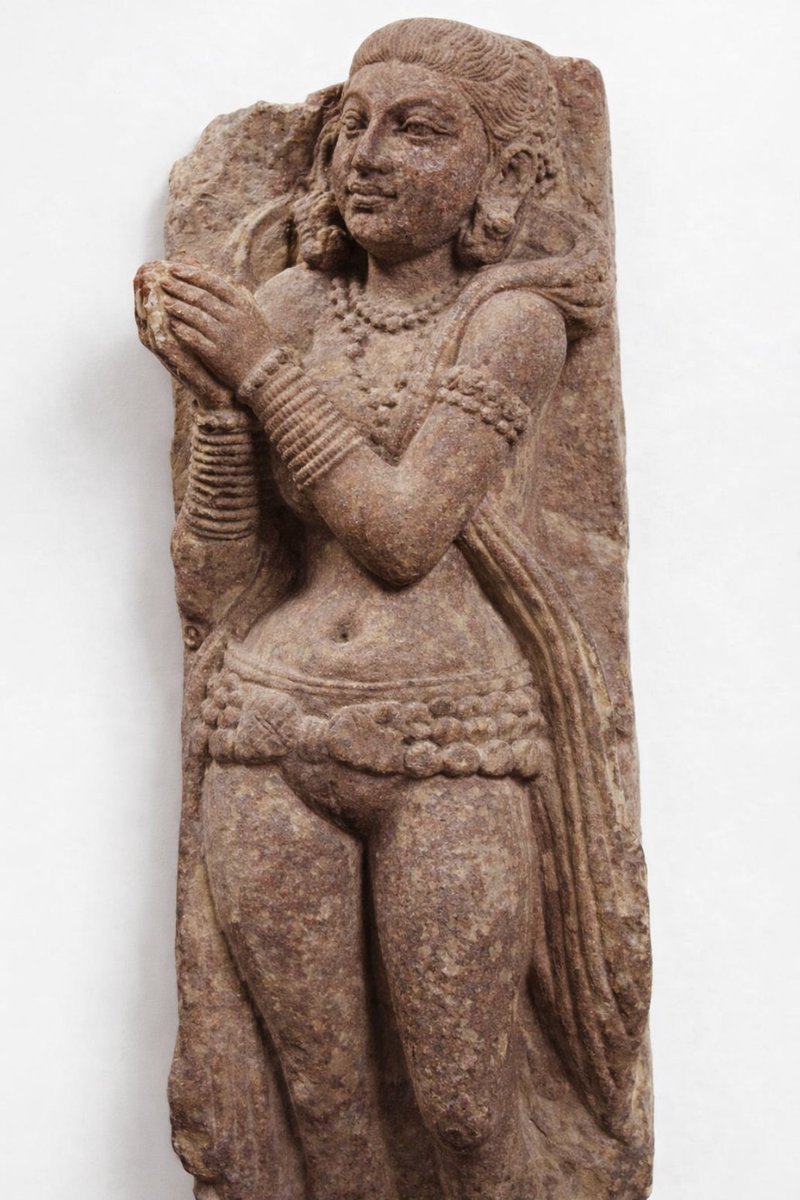 Mathura 200 CE

She does not shout power.
She folds it.

Sandstone. Red. Weathered. Still breathing.

Hands in prayer. Not submission — composure.
Anjalī is geometry. A vertical axis. A social signal.

Look at the uttariya.
Not stitched. Not tailored. Draped. Intelligent cloth.