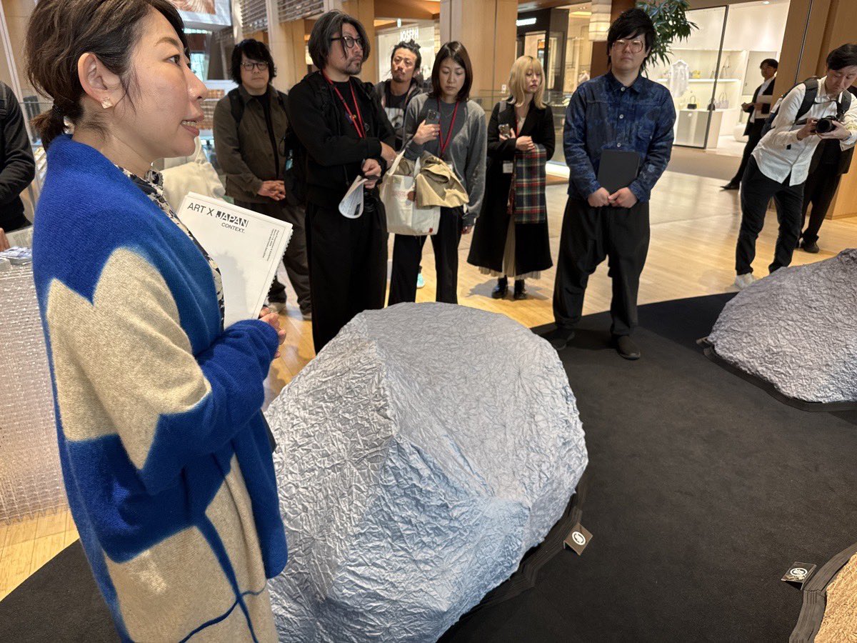 nobi's tweet photo. #ARTxJAPANCONTEXT

「ART X JAPAN CONTEXT.」最終作品発表・展示会
各作品紹介【前編】

* **松山周平×スズサン：「Encoded Nature on Cloth」**
* スマートフォンで3Dスキャンした岩のデータを基に、ぬいぐるみを製作。絞り染めの職人との協業により、3Dプリントによる構造体と手作りのテクスチャーを組み合わせた作品である。
* テクスチャーの歪みを抑え、絞り職人が作業しやすいデータに変換する専用プラグインソフトウェアを開発。オーストリアの氷河からの石（2億年前）と神奈川県の海辺の岩（20万年前）をモチーフに、自然素材を使わずに「岩らしさ」を再現し、自然の本質を問いかける作品である。
* 3Dスキャンにはアプリストアの「スリーディー」という骨格アプリを使用。また、絞り技術は有馬地区で伝統的に綿や麻などの天然繊維を染める技術であるが、本作品ではポリエステル素材に応用し、形状記憶を付与する新しい技術が用いられている。これは通常、照明器具などに使われる技術の立体作品への応用である。

* **涌井智仁×Design Solution Forum：「アトラスズ」**
* 半導体とビジュアル表現の関係性を探求するリサーチベースの手法であり、インスタレーションや映像で展開される。
* 冷蔵庫をサーバー運用やデータ収集のための最適化された箱として再解釈。デジタルデータが20〜30年で消失する問題に取り組み、ハードディスクに負荷をかけ続け、世界中のオムレツレシピ（現在517枚）を常に収集している。
* エラーを検知すると信号が赤になるダイアグラム的な仕組みと、リサーチ内容に基づく映像作品を展示する。

* **snipe1×セーレン：「BOM（b）MATSURI（まつりだぼん）～UNIVERSE FLOWS NOT CIRCLE BUT SPIRAL～」**
* 新しい盆踊りをテーマに、伝統文化と現代アートを融合したプロジェクトである。
* 「ドロップ」（ストリート用語で新しいものを爆発的に発表する意）をコンセプトに、和紙との協業や文化祭形式で実施。校内放送や渡り廊下でのインスタレーション、研究者による「人はなぜ回るのか」というテーマの講演も実施された。
* 六面体の新しい形状を制作し、その周りを回る体験を提供する。

* **杉浦久子×東洋竹工：「市中の山居―橋上の茶室 WA-AN（和庵）、東京ミッドタウン」**
* トランク一つから展開できる可搬式の茶室作品である。
* 「ナイキン玉垂れ」という、重力に従った自然な曲線美を表現する技法を使用。東京、新潟、テヘラン、パリを巡回し、今回は公共スペース（往来）の真ん中に、橋の上に置いたようなイメージで展示される。
* 利休の待庵の平面図を参照し、にじり口（身をかがめて入る小さな入口）のコンセプトをリスペクト。1日5回、各回2名のゲストを招いてお茶会を実施する。
* テヘランで展示した作品は持ち帰れなくなり、今回新しく制作されたものである。

* **井村一登×ExtraBold：「bulbocodium」**
* カラヴァッジョの絵画「ナルキッソス」をモチーフに、巨大3Dプリンター会社エクストラボルトと共同制作された作品である。
* ナルキッソスの自己愛を肯定する試みとして制作され、珍しい自家受粉する水仙「ナルキッソス・バルボコディウム」が配置されている。
* 作者自身の結婚経験から、他者と苦しみや喜びを共有できないナルキッソスを肯定したいという思いが込められている。作品の目には小型カメラ、頭部にはパソコンが内蔵され、胸元から放熱しており、小型カメラで捉えた景色を水仙のモジュールに投影することで、ナルキッソスが見ていた景色を主観的・客観的に体験できる。

(つづく)