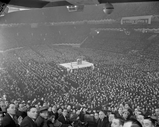 20thCentBoxIMM's tweet photo. It's a packed house at Madison Square Garden, on March 7, 1938, for the finals of the New York Daily News Golden Gloves Tournament.

The New York Times writes:

"Twenty thousand boxing fans will pack Madison Square Garden tonight to witness the finals of the Golden Gloves-New York's leading amateur program of the year."

From a two-part Boxing News Online article from 2020 on the rise and fall (and, hopefully, someday the rise once more) of The New York Daily News Golden Gloves tournament (I'll post Part II of this article in the comments section):

IN court they call it irreconcilable differences, which is the inability of the two parties to resolve their problems. In a nutshell, that explains why one of boxing’s great institutions, the New York Daily News Golden Gloves amateur boxing tournament, is no more. This is a story of how greed, ego, power, and people’s inability to communicate destroyed a long and honoured tradition. This is a story of how dreams were shattered because the people in authority were more interested in serving their own interests than the athletes they were entrusted to look after.
The Daily News is a New York City newspaper that is now over 100 years old and at one time had a circulation of over two-million readers. In 1927, DN sports editor Paul Gallico started what became known as the Golden Gloves. It fast became a tradition that transcended boxing. Officially it has always been called the Daily News Golden Gloves; The tournament run by New York News Charities Inc., with the profits being given to various causes.
Those who participated over the years came from diverse backgrounds. The cultural phenomenon represented the best of New York City. The Daily News’ vast circulation not only read about the fights, but the stories of the fighters as well. The more they won the better known they became to the public. The boxers fought for glory, the right to be called a GG champion, and to have a small pair of Golden Gloves placed around their neck. Those pair of gloves were as precious to them as a gold medal was to an Olympic athlete.
How important it was to some can be found in the story of super-heavyweight Angel Alston. After losing in the finals of the 1984 tournament, in his first try at the Gloves, Alston kept chasing his dream unsuccessfully for the next 10 years. Finally on his last year of eligibility before age requirements would end his career as a Golden Glover, Alston broke through. As they say, you can’t make this stuff up.
Becoming a Golden Glove champion was a life-altering experience. At a time when neighbourhood ties were stronger than they are today it meant instant celebrity status within that patch of land. Winning the GGs was the ultimate achievement as so many of the alumni attest to. No matter what they achieved afterward, the Golden Gloves always held the most special of places in their heart. Mark Breland, who won the Gloves five consecutive years (1980-1984) and is associated with the tournament more than any other boxer in its history, vouches for that. “It means far more to me than my Olympic gold medal and two world titles as a pro,” he said.
Vito Antuofermo who won the Gloves in 1970 then later held the undisputed world middleweight championship, concurs. “If I could choose only one, the Golden Gloves would be it,” he said. “It is the greatest thing I ever experienced.”
The New York Daily News Golden Gloves was the springboard for great fighters who would go on to Hall of Fame careers as professionals, a list that includes Sugar Ray Robinson, Floyd Patterson, Emile Griffith, Hector Camacho and Riddick Bowe. Those names are legendary, but the Golden Gloves was never regarded as a developmental league to launch professional careers. Public perception withstanding, to those who won it the NYGG was the pinnacle, the highlight of their youth so to speak. And the memories were not only confined to those doing the punching. It was a place where family and friends bonded in support of their favourites. It was a tournament that built character, got kids off the streets and propelled them to an improved lifestyle they might not have been able to find elsewhere.
“These are not the kids who will get in trouble, these are the kids who are trying to do the right thing,” then-New York State Senator Al D’Amato told me at ringside during the night of the 1988 finals. The evidence backed that up. In an era when drug use among young people was rampant, the Daily News, for the first time, had invoked mandatory drug testing for the tournament. When all was said and done of the 744 who were tested only six were positive – less than one percent.
For the first 60 years of the tournament the finals were held at Madison Square Garden, then as now the world’s most famous arena.
Garden jitters had been a term frequently used for boxers who under-performed in the big house. Certainly the pressure of appearing in the Gloves finals was something that was impossible to prepare for. The wait in the dressing room to be called was nerve racking. Then the moment of truth arrived, where the boxer made the walk through the tunnel, down the aisle, and into the ring where a sudden silence would ensue. The lights in the Garden would dim, then the spotlight would shine directly on the boxer as he was introduced to the packed house telling us the gym they represented, their tournament record, and where they went to school or were employed. The fighters nervously braced for those introductions as they would the sudden dip of a roller coaster, the intensity as unbearable for some and as it was uplifting for others.
Three rounds to justify the hard training and sacrifices made, the mornings on the wintery road, the nights at the gym. Some of the championship contests produced a clear winner, others ended in nerve-racking suspense where the final bell rang with the outcome in doubt. The two fighters would gather beside the referee in the centre of the ring where they would await the decision that would impact them for the rest of their life. For the loser the effort was secondary, the pat on the back telling him good try was of little consolation. Getting to the top of Mount Rushmore is so much better than saying you had a great view of it, after all. The fight was over but the adrenaline continued to run in the few seconds before they would learn their fate. It was not uncommon for many to try to will their way to victory. They hoped, prayed, anything that would tilt the scorecards their way.
It lasts literally seconds, just as a jury verdict does. “The winner in the — division is —“. The pure ecstasy of being declared new champion was indescribable. They had joined an exclusive club and would always have a title next to their name. That moment would be vivid in their mind for the rest of their life. One they would happily hold onto and keep reliving. Unless they had won the Gloves before or would do so in a subsequent year, those who did not hear their name announced at the end would always wonder ‘what if’.
The journey began by filing out and mailing in the application form that would appear in the Daily News sports section. For some the planning was meticulous, having spent time in the gym under the watchful eye of a coach where they gained experience and a high level of fitness. Then there were those who had never boxed before and had an inflated image of their prowess: They would sign-up on a whim, take a crash course at their local gym, then learn the painful realities of the manly art of self-defence, never to return to the tournament again.
The early matchups in the tournament were not created by coincidence, but by design. Even in the so-called dark ages where cell phones and the internet would have been an invention beyond the imagination, there were pretty good scouting reports on the better boxers entering the Gloves. The competition at unsanctioned events meant that in some instances boxers might have already squared off before facing the other in Golden Glove competition. The seeding, therefore, would pair the best in each division against the presumed weakest in the hopes of saving the marquee matchups for last. It was a formula which worked and quickly distinguished the pretenders from the contenders.
The good times, at least those we knew way back when, would not last forever.
When less than 12,000 turned out for the 1987 finals at Madison Square Garden, it marked the end of an era where the championship matches would be contested in the main arena. MSG continued to host the finals, but they now took place in its smaller venue, the Felt Forum, now known as the Hulu Theater. It was more than the crowd size that factored into the decision. The number of weight divisions was increased meaning that the number of fights made for too long of an evening. Beginning in 1988, the finals would take place over the course of two nights, which MSG would host until 2012.
The decline, I suppose, was inevitable. Even so, it should never have been allowed to plummet to the inescapable depths it now finds itself.
No one particular person is responsible for the demise of the Daily News New York Golden Gloves, but it becomes apparent after investigating the tug of war that went on behind the scenes that a toxic atmosphere existed between the Daily News Charities Inc; and Metros in which coaches were said to be intimidated and forced to choose sides. A mini-war escalated in which the prospective Golden Glovers were the ultimate victims.
Historically, the Golden Gloves had always mirrored the geographical makeup of New York City. Over the decades the diverse ethnicity of the tournament mirrored life in the town. But as times changed so did the number of entrants, less of them signing up to compete in the Golden Gloves.
At one time it was the exact opposite. In 1937 at Madison Square Garden, three rings simultaneously held the entire quarter-finals of the tournament over one day where an astounding 107 matches occurred. But as we moved into the following century, inequities existed that were never properly addressed. As journalist Ryan Songolia, who covered the tournament for the Daily News in 2011 pointed out, “Some weight divisions had less entrants than others making the road to the finals much easier. Some fighters had to box only once to get there because of a lack of competition in their division. Others had to box a few times at least.”  This was especially true when women’s boxing became a part of the tournament in 1995 and did not have nearly as many participants as did the men.
Smoker shows held in churches, gyms, and youth centres in the various neighbourhoods of New York are a thing of the past, but at one time they were very popular on the amateur circuit. In fact, in the early 1980s, Mike Tyson travelled several times from the Catskills to the Bronx, to box on them. These unsanctioned shows fought in front of boisterous crowds gave the boxer invaluable experience entering the Gloves tournament. In a sense it made a mockery of the GG Sub Novice category that was intended for participants who had no prior fights at the time. Tightened insurance laws ultimately made putting on these shows cost prohibitive.
What is indisputable is the impact that the New York Daily News had on the tournament during its peak years. As the newspaper with the largest circulation in New York City, the Daily News took special pride in the tournament being theirs. The coverage they provided of all the shows from beginning to end was extensive. Come tournament time the back page of the paper would routinely have photos of the action the evening before.
As other sports gained in popularity, the New York Golden Gloves was sure to suffer, but amateur boxing had an unexpected revival with the charismatic 1976 United States Olympic team anchored by Sugar Ray Leonard and Howard Davis Jnr. The five gold medals at those Montreal games captured the imagination of the American public. Davis Jnr from Glen Cove, Long Island, was a four-time New York Gloves champion (1973-1976) and it was he who won the Val Barker trophy as the outstanding boxer of those Olympics. In the same year, the Academy Award-winning movie Rocky inspired a new generation of amateur boxers as well.
When Mark Breland entered the picture a few years later, the New York Golden Gloves had their most charismatic entrant in the tournament’s history. Breland regularly scored highlight reel knockouts and attendance soared on the nights he boxed. But Breland was the exception to the rule, the rare fighter who could attract fans to shows for the sole purpose of seeing him box. Over the years it had transitioned to the point where, with the exception of the finals, the only ones attending the events were family and friends of the boxers. In addition, with attendance being down the once vibrant atmosphere was gone. On many occasions people left the moment the match ended they had come to see.
The NFL’s popularity skyrocketed in the 1970’s and the NBA’s in the mid-eighties. As wildly popular as the NBA is today, it was not even a primetime sport in 1980. In fact, as incredible as it seems now, the decisive game of that year’s finals, which saw Magic Johnson’s Los Angeles Lakers defeat Julius Erving’s Philadelphia 76ers, was not even shown on live television.  As the landscape changed and increased attention was given to other sports, the Golden Gloves was not as prominently displayed on the pages of the Daily News as before.
Golden Gloves fans had long been a devoted group braving the winter elements to attend shows, but when cable television came into existence with its broad array of programming the motivation to head out the door was compromised. But what hurt more was the advent of the internet, something that has proven to be a crippling blow to newspapers and publishing industries around the globe. Staff has been cut and jobs lost for them to stay in business. These days, you can obtain virtually any information, albeit of varying quality, by going onto the internet. Hit hard by this, the New York Daily News was no longer the major media player it had once been.
The New York Daily News Golden Gloves was the first of its kind, but many other cities followed in having tournaments of their own, many enjoying success. The Chicago Golden Gloves was popular for a time. There would be inter-city rivalries with The Big Apple in the 1950s through the mid-60s, then resume for a few more years from 1977 to 1982. When New York hosted one such event at Madison Square Garden in 1960, among the winners for the victorious Chicago team was one Cassius Marcellus Clay. In 1983, when the Chicago Tribune withdrew their sponsorship the inter-city rivalry was done, but for those who were around at the time it conjures up great memories.
For all the success that other tournaments had there was no disputing that the New York Daily News Golden Gloves stood above the rest. It was the one tournament where amateur boxers from all over came to be a part of. Among them was Tony Madigan, the British Empire and Australian 175lb champion, and an Olympian in both 1952 and 1956. Madigan travelled the world for the sole purpose of winning the New York Gloves. He accomplished his mission in the 1959 tournament, but would later lose to Clay in the inter-city competition fought in Chicago. Clay who later became known as Muhammad Ali, continued to be a nemesis to Madigan, defeating him in the 1960 Olympics in Rome as well. Those heady days of Madigan and Clay being associated with the Gloves is now a memory so distant it’s scarcely believable.
The problems between the New York Daily News Charities Inc; and the USA Metro had been building up for some time until it came to a head in 2017 which was the last year of the NY Golden Gloves. USA Boxing’s position was that the tournament was being run poorly and that they could do a much better job of serving the participants. They were also of the opinion that the Daily News charities were making a significant profit which should have been used in part to finance boxers and their coaches for out of town trips. According to people involved with the DNC, the tournament had ceased to become profitable due to high operating costs and diminished attendance at events.
The issues with how the DNC should distribute potential profits dates back to 1947 when the Amateur Boxing Coaches Organization (ABCO) made several demands that were rejected. ABCO then unsuccessfully tried to get the boxers to boycott the tournament. The Metropolitan AAU, the sanctioning body of the time, moved quickly and decisively on the side of the DNC putting the matter to rest. Such was the clout that the New York Daily News had in its heyday. But this is an altogether different era, and standing directly in the line of fire was Brian Adams, who beginning in 2006 was entrusted with running the tournament by the DNC (Daily News Corp.).