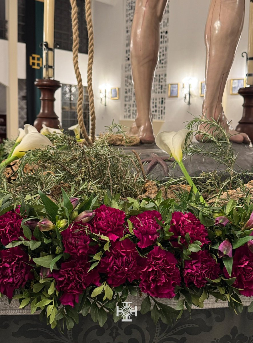 La mesa de las andas simula un pequeño monte natural, sobre el que se dispone un exorno floral compuesto por claveles y astromelias en tonos morados, verdes de lentisco y romero, salpicado con calas blancas, dispuesto por Floristería Rose.