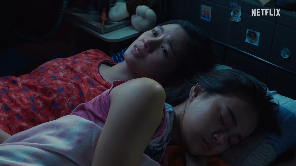 A still from a Netflix film showing a woman lying awake next to a sleeping girl in a dimly lit bedroom.