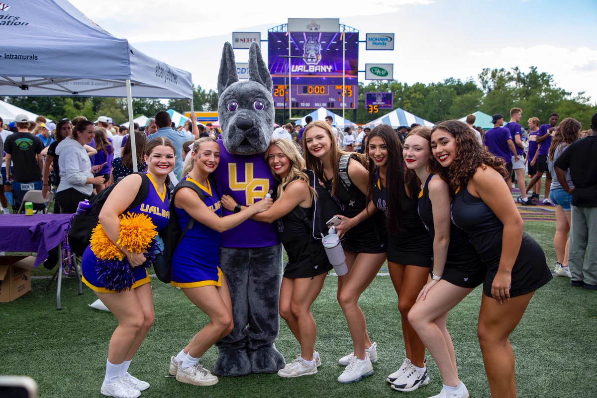 A group of people is standing on a sports field with the UAlbany mascot. They are posing for a photo with a scoreboard showing UAlbany's logo in the background. Other attendees and tents are visible in the surrounding area.