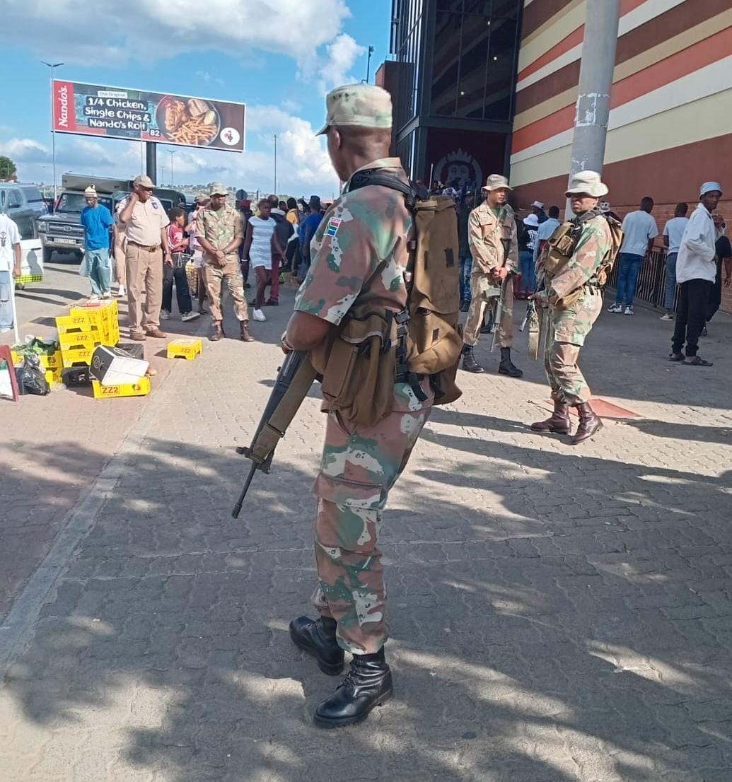 South African National Defence Force soldiers deployed in an urban area, standing armed on a street with crowds, market stalls, and mountains in the background under a partly cloudy sky
