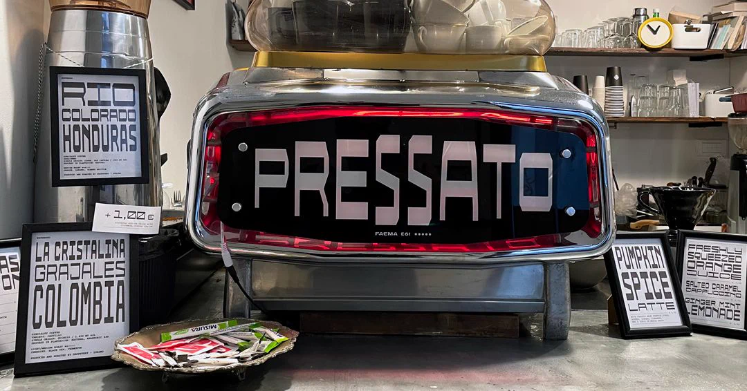 A coffee machine with large white letters reading "PRESSATO" is prominently displayed on a counter. Surrounding the machine are various framed signs. Coffee-making equipment and small decor items are visible in the background.