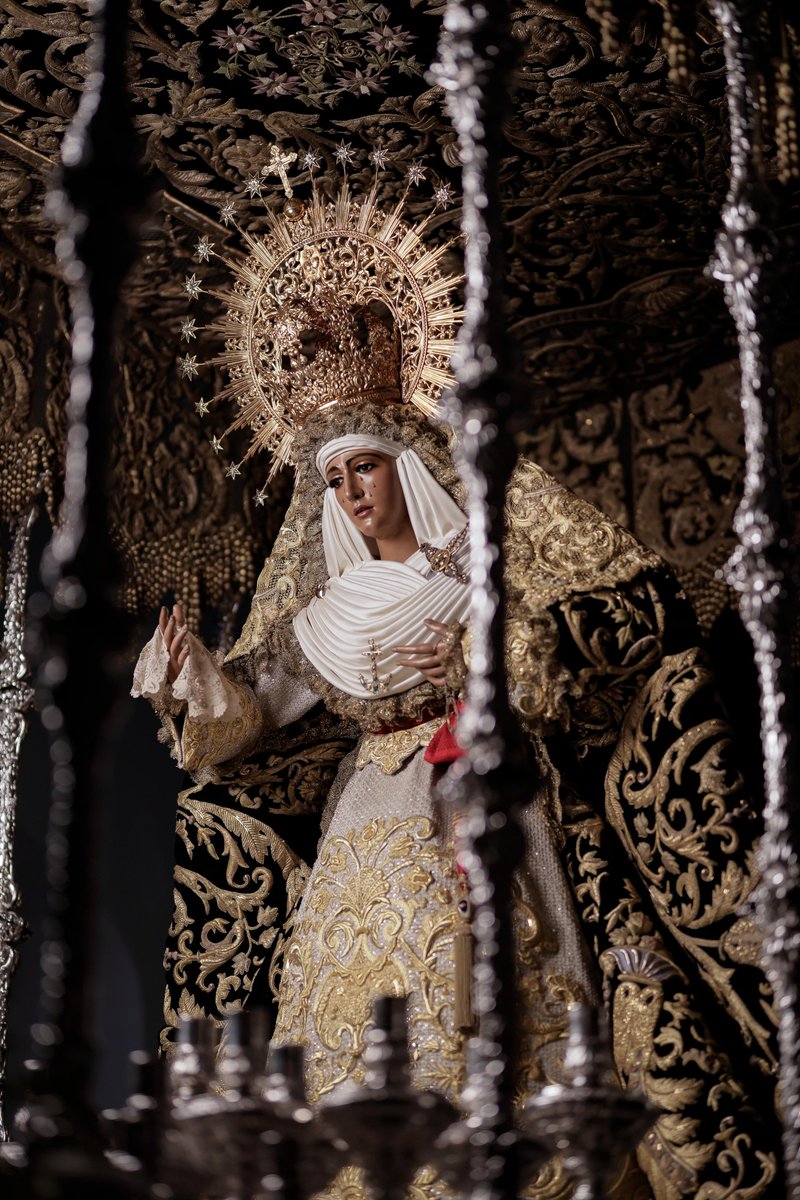 EspDeTriana's tweet photo. Nuestra amantísima titular luce sobre sus benditas sienes la nueva toca de sobremanto ejecutada también en el taller de Manuel Solano, bajo el diseño de Gonzalo Navarro; de inspiración plenamente barroca. 

Centra el conjunto de la misma una jarra de azucenas sobre la que se sitúa una cartela que alberga una representación de la Asunción de Nuestra Señora, en clara alusión al papel desempeñado por la hermandad en defensa del Dogma, del que el pasado año 2025 se celebró el setenta y cinco aniversario de su proclamación.

En esta ocasión, la Santísima Virgen porta el popular manto “de los dragones”, realizado por el taller de los Sobrinos de José Caro, según diseño del ceramista José Recio del Rivero. Está bordado en oro y sedas sobre terciopelo verde oscuro. Es considerado como una de las joyas de la Semana Santa Sevillana, tanto por su maravilloso dibujo, como por la perfección técnica que Esperanza Elena Caro aplicó en él, causa que hizo que lo definiera siempre como una de sus mejores obras. En él vemos un amplio muestrario de la mayoría de los puntos utilizados por tan genial maestra del bordado.

El rostro de nuestra amantísima titular se enmarca en un bello tocado de raso de seda, enjoyado con el puñal y el ancla del siempre recordado Fernando Morillo.

En la cintura de la Esperanza se ciñe el fajín de Capitán General de la Armada de S.A.R. Don Juan de Borbón y Battenberg, donado y entregado personalmente, en una ceremonia celebrada el 29 de Septiembre de 1998, por su Camarera Honoraria Perpetua desde 1958, S.A.R. Doña María de las Mercedes de Borbón y Orleáns, en nombre de S.M. el Rey de España Don Juan Carlos I.

El conjunto se culmina con la presea que corona las benditas sienes de Nuestra Señora de la Esperanza, realizada en 1937 por el orfebre Gabriel Medina, para cuya ejecución se reutilizó el canasto de una anterior, la realizada por el platero Justino de Guzmán entre 1890 y 1891.