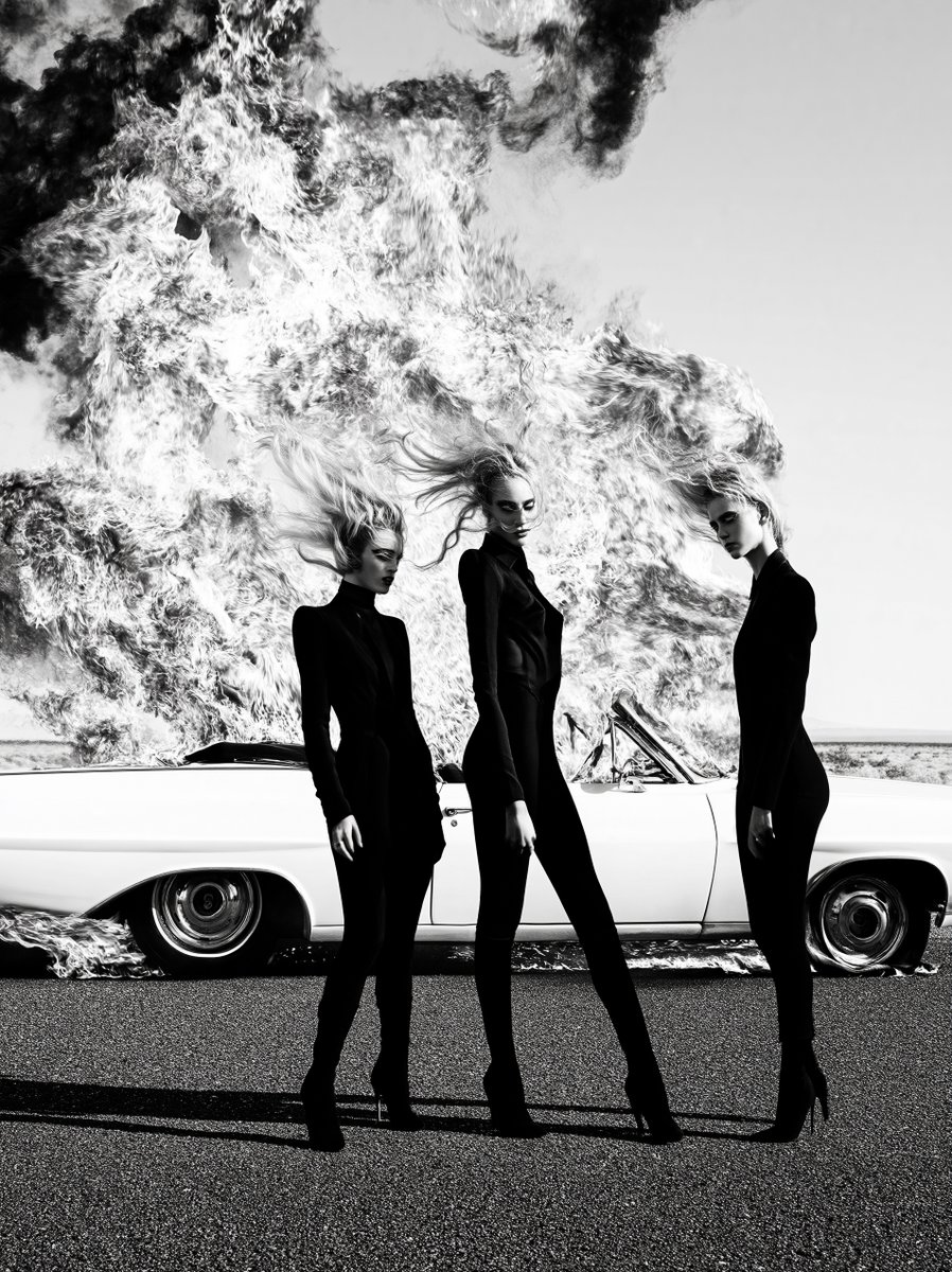Black and white cinematic fashion photograph of three women standing in front of a burning vintage convertible in the open desert. towering hair. fitted black silhouettes. hard wind. low angle composition. flames rising behind them in a sculptural wall. chrome reflecting firelight. rigid poses. sharp eyeliner. iconic tableau. cult cinema intensity. luxury editorial rendering. immaculate monochrome contrast. unforgettable still image --ar 3:4 --sref 2298369550 --profile oovu5h7 --hd