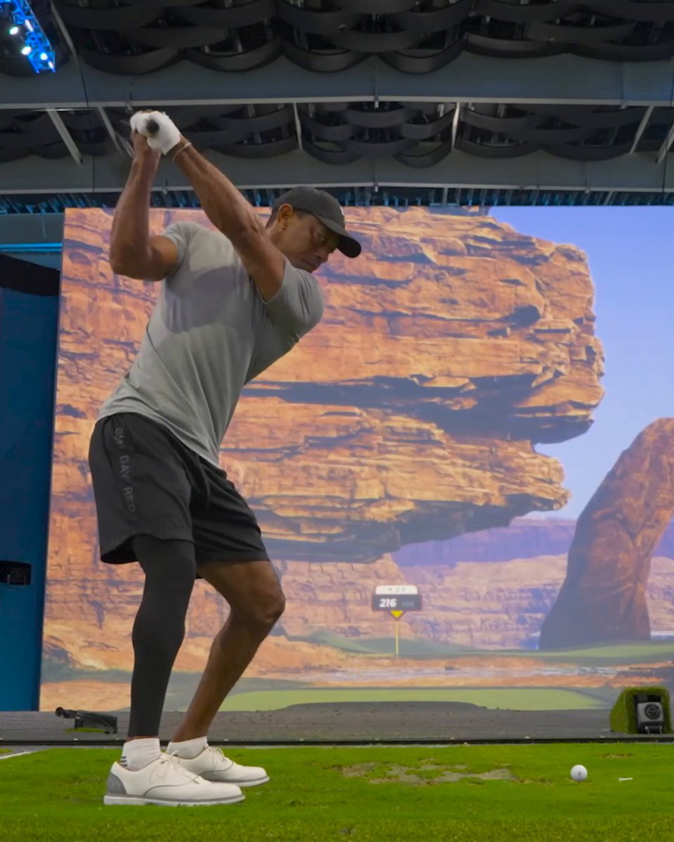 A golfer in a gray shirt and "OUR DAY RECO" shorts completes his swing at an indoor golf simulator, with a screen displaying a desert landscape and "216 YDS" to the pin. The post "Give them one of these" humorously suggests the powerful swing is worthy of receiving a "one of these" — likely referencing the impressive distance displayed on the virtual course.