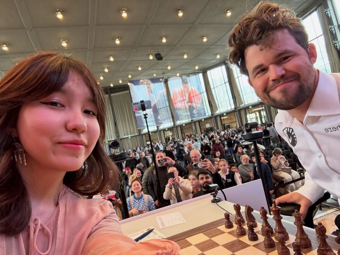 chess1square's tweet photo. صباح الخير 

تجمع هذه الصورة بين أسطورة الشطرنج والمصنف الأول عالميًا **ماغنوس كارلسن (Magnus Carlsen)**، واللاعبة الكازاخستانية الشابة الحاصلة على لقب أستاذة دولية كبيرة **ألووا نورمان (Alua Nurman)**.
التقطت الصورة في أوائل **أبريل 2026** خلال بطولة "غرينكه تشيس فري ستايل المفتوحة" (Grenke Chess Freestyle Open) في ألمانيا، وتحمل وراءها قصة طريفة انتشرت بشكل واسع مؤخرًا:
 * **طلب عفوي:** قبل بدء الجولة الثانية من البطولة، وجدت اللاعبة الشابة ألووا (18 عامًا) نفسها في مواجهة كارلسن على الطاولة رقم 1. ومن شدة حماسها للعب أمام المصنف الأول عالمياً، طلبت منه التقاط صورة "سيلفي" للذكرى.
 * **موافقة ثم صرامة:** وافق كارلسن بلطف وابتسم للكاميرا (كما هو واضح في الصورة). ولكن، بمجرد أن انتهت من التصوير، قام كارلسن بهدوء بتبليغ حكم البطولة بوجود هاتف محمول مع منافسته.
 * **تطبيق القواعد:** تمنع قواعد الاتحاد الدولي للشطرنج (FIDE) الصارمة لمكافحة الغش اصطحاب الهواتف المحمولة أو أي أجهزة إلكترونية إلى طاولة اللعب تمامًا. وبناءً على ذلك، جاء الحكم وصادر هاتف ألووا حتى يتسنى لهما بدء المباراة بشكل قانوني.
**خاتمة الموقف:**
انتهت المباراة بفوز ماغنوس كارلسن بعد 44 نقلة، لكن اللقطة أثارت تفاعلًا كبيرًا على الإنترنت؛ حيث أشاد خبراء اللعبة باحترافية كارلسن وصرامته في تطبيق القواعد لحماية نزاهة البطولة، بينما رأى آخرون أن الموقف كان محرجًا وطريفًا بسبب التحول السريع لكارلسن من ابتسامة الـ"سيلفي" إلى الإبلاغ الفوري للحكم!