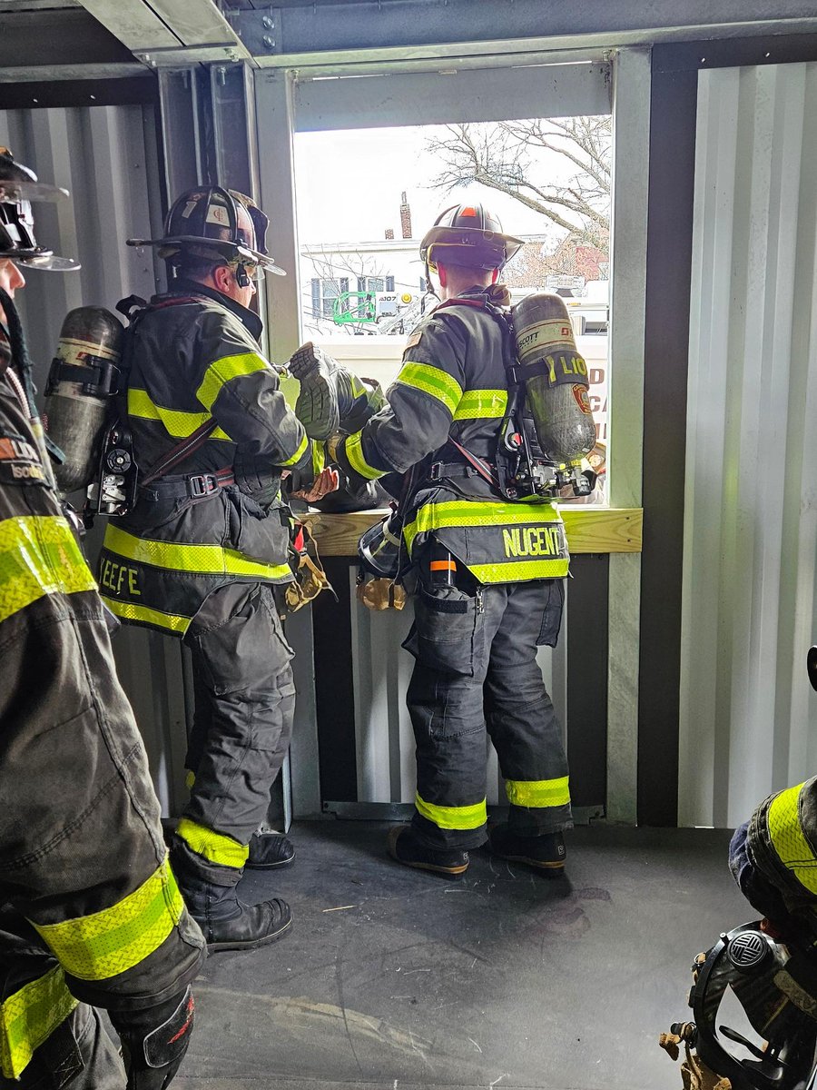 Firefighters in full gear are training in a metal structure. One is using a tool at a window, while others observe. Trees and buildings are visible through the window.