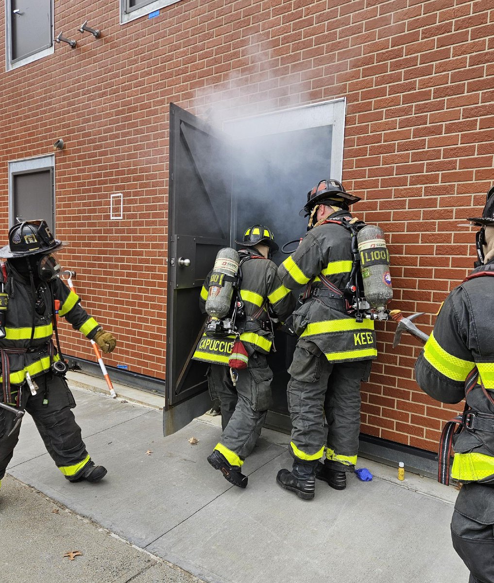 Firefighters in protective gear are entering a smoke-filled doorway of a brick building. They are wearing self contained breathing apparatus and helmets.