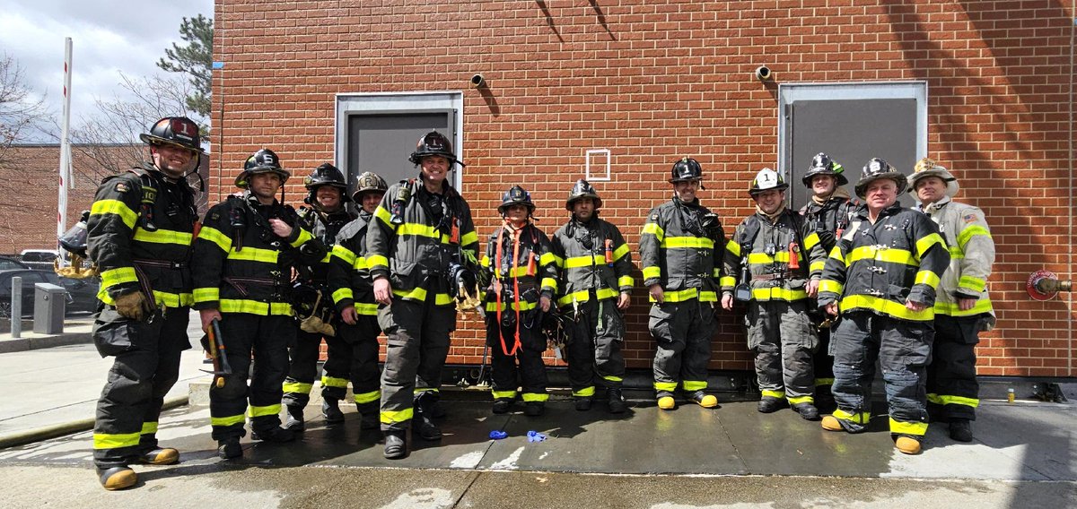 A group of firefighters in full gear stand together in front of a brick building.