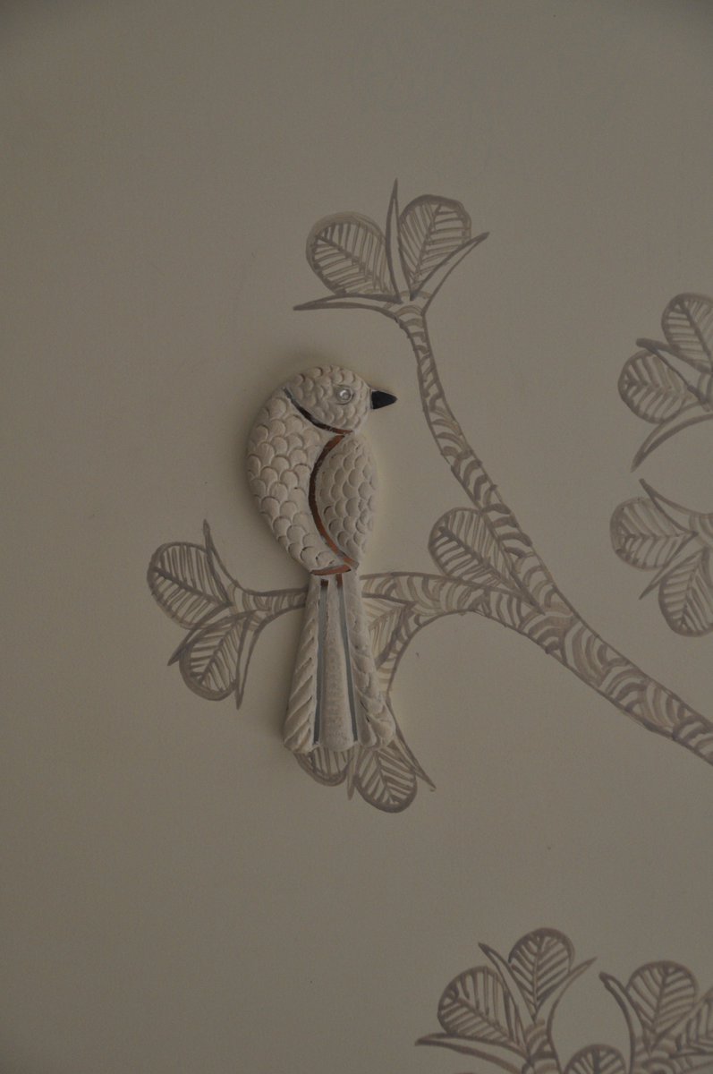 CraftCanvas's tweet photo. In 2012 (I think), we got a brief for a living room mural. It had to be subtle and something that they can live with for years to come. It had to have the same sense of calm that the owners did, absolutely trusting us and the process.
It was our first attempt at combining two crafts in one installation. We didn't know then that it would start off a path for us.
The wall is in Gond painting, the tradition of the Gondi people of central India, where every line is the logic of nature, every form connected to every other form. Painted in silver-grey on a warm white wall. No strong colour, just the suggestion of a forest.
Into that forest we placed birds. Three-dimensional. Made in mud mirror work which is a craft tradition where clay is shaped, dried, and inlaid with tiny mirrors. Each bird perched on a painted branch or is flying towards one, one foot in the two-dimensional world of the Gond painting, one foot in the physical world of the room.
The palette was deliberately muted as the brief demanded it. But the glass shards in the mud mirror work meant the installation was never the same twice. Morning light read it one way. Afternoon another. The birds caught what the wall absorbed.
Two crafts. Two dimensions. One forest that changed with the light.
We have never been able to fully explain why this combination worked as well as it did. Some things you discover by doing, not by planning.
Team: Artisan Kaushal Prasad Tekam, Surprit Dang
Made in collaboration with IDO DESIGNS