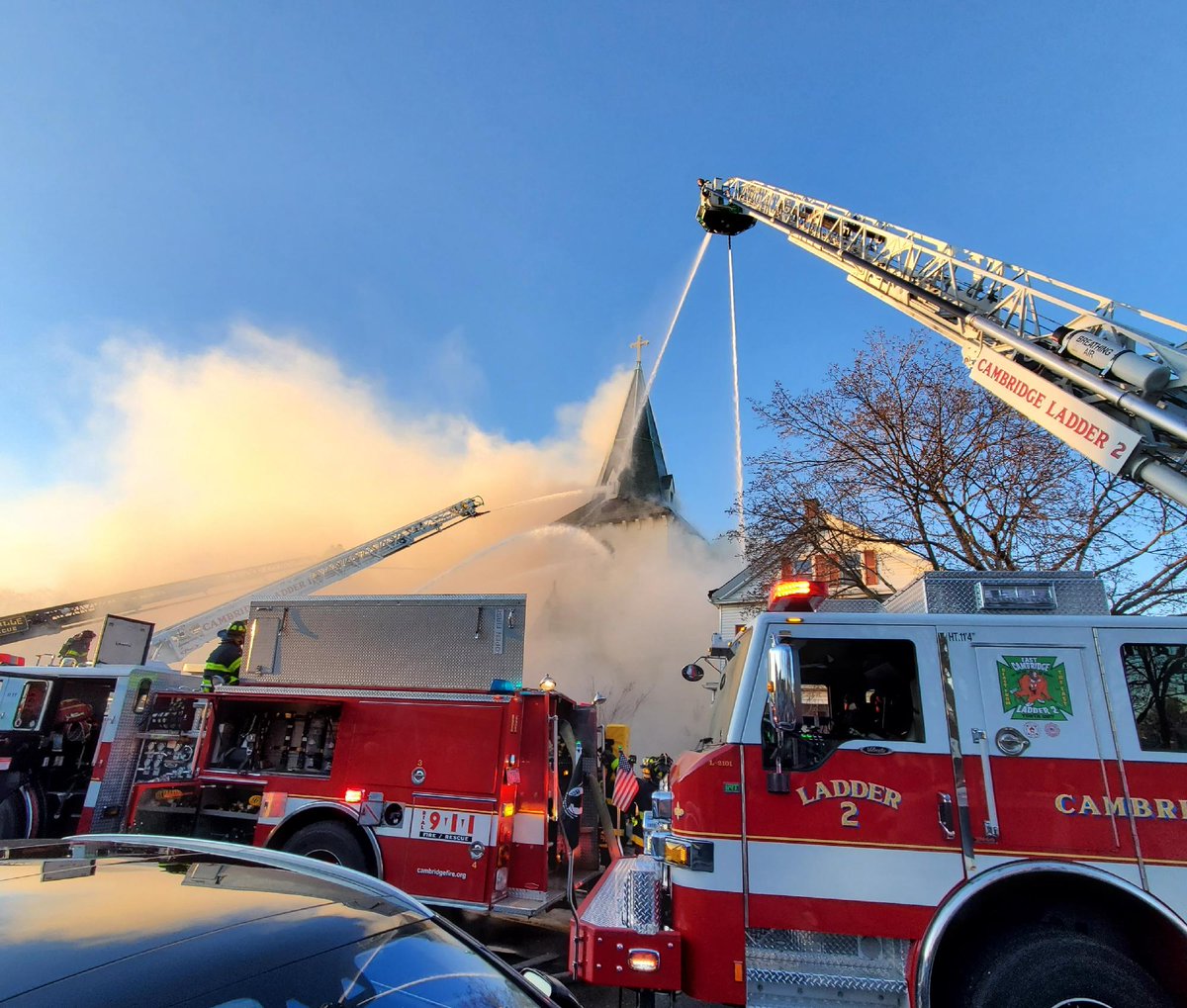 Firefighters respond to a large fire at a church with a prominent steeple. Thick smoke billows into the sky. Several fire trucks, including ones labeled "Cambridge Ladder 2," are positioned on the street, using ladders to reach the flames. Crowds and emergency responders are visible nearby.