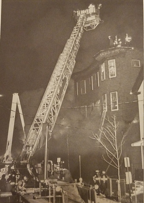 A large ladder extends from a fire truck to the roof of a multi-story building that is emitting smoke. Firefighters are positioned on the roof and around the building. The scene is illuminated by lights from the emergency vehicles, and a crowd is gathered at a safe distance. It's nighttime, and the atmosphere is tense.