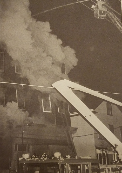 Firefighters respond to a burning multistory building at night, using a ladder truck to direct water onto the fire. Smoke billows from the windows as they work to extinguish the blaze.