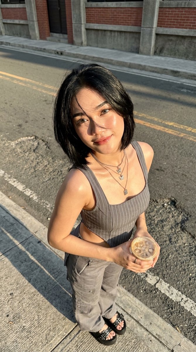 Ciahasyim's tweet photo. Prompt

A stunning young Southeast Asian woman in her early 20s, sharp Indonesian features, glowing tanned skin with a natural dewy sheen from the sun, high cheekbones, sultry dark almond eyes with subtle eyeliner, full pouty lips with a soft confident smirk, perfectly shaped eyebrows, short jet-black bob haircut just past chin length, windswept strands dramatically blowing across her face, one lock teasing over her right eye, looking straight at viewer with playful bedroom eyes.
She’s standing on a cracked concrete sidewalk next to an asphalt road with faded yellow lines, body slightly twisted toward camera, weight on one leg, midriff exposed, wearing a tight gray pinstripe sleeveless crop top with thick shoulder straps and square neckline that hugs her perky cleavage, matching low-rise gray cargo pants with visible zip pockets and utility details, black chunky platform sandals with multiple straps and silver buckles.
Layered silver necklaces dripping down her chest — one with tiny butterfly pendants, another with a square charm — delicate ring on her left ring finger. Both hands gently cradling a clear plastic cup of iced coffee with visible ice cubes and condensation, straw ready.
Golden hour sunlight hitting from the side, strong warm highlights on her skin and hair, dramatic long shadows on the ground, realistic skin texture with light sweat glow, photorealistic, ultra-detailed 8k, cinematic street photography style, natural color grading, sharp focus, shot from a slightly high angle, urban brick building with concrete pillars in soft background, sunny afternoon vibe, confident street fashion mood, seductive yet casual energy, maximum detail, no blur, masterpiece.
C. Hasyim