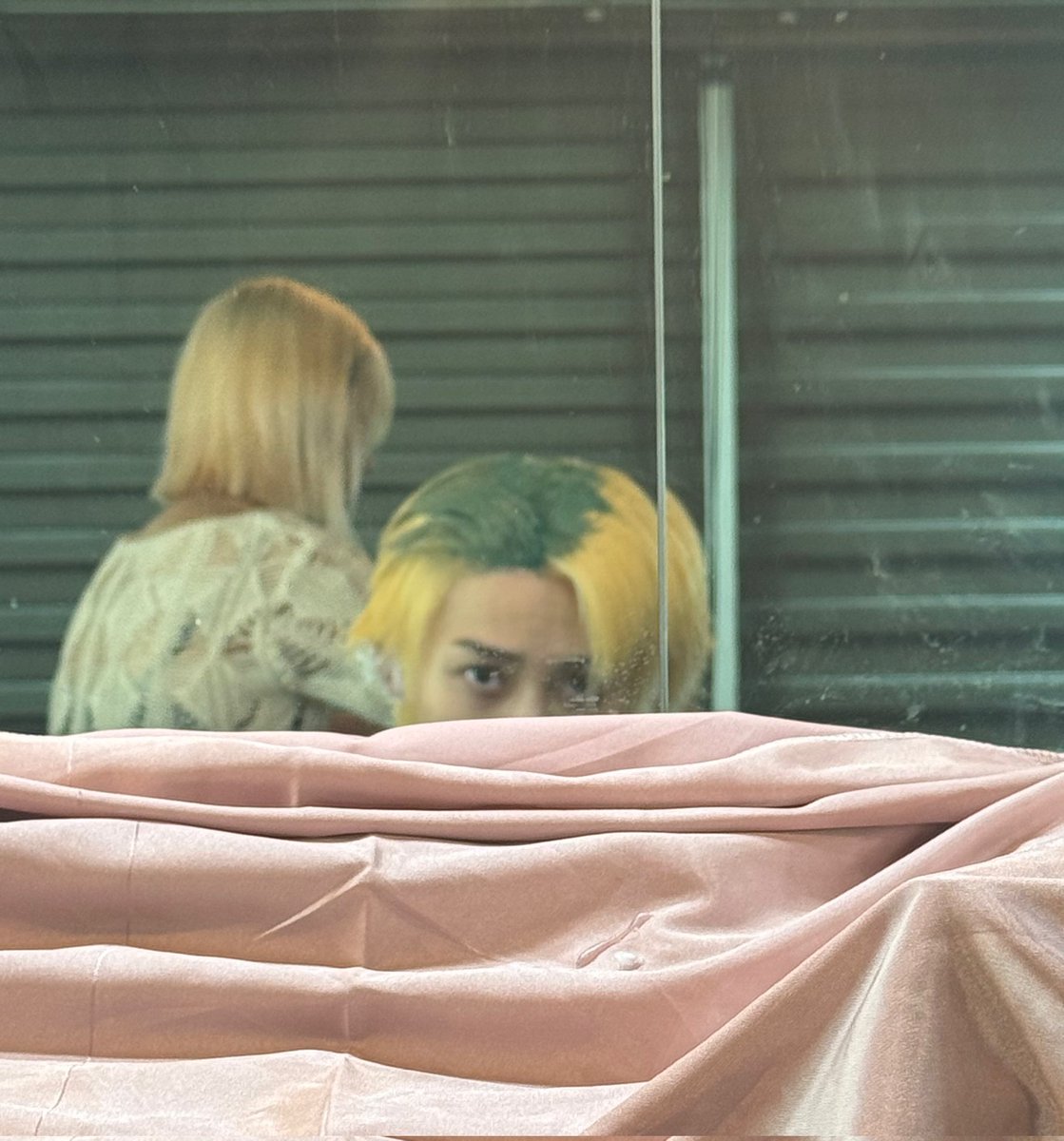 A muted room framed by glass, where a figure with two-toned hair peers just above soft pink fabric. The mirror doubles the scene, adding distance and quiet tension.