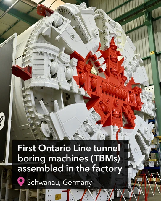 A construction site featuring a large tunnel boring machine positioned in the centre. Surrounding the machine are high-rise buildings and a crane. The CN Tower is visible in the background against a blue sky with clouds. Text over the photo reads "First Ontario Line tunnel boring machine (TBM) assembled in the factory".