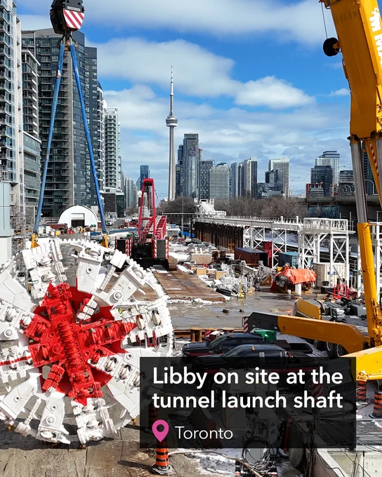 A large tunnel boring machine is displayed inside a green-roofed industrial building. The central part of the machine is distinctively marked with a red star-like design, while the outer section is white. Text over the image reads "Libby on site at the tunnel launch shaft".