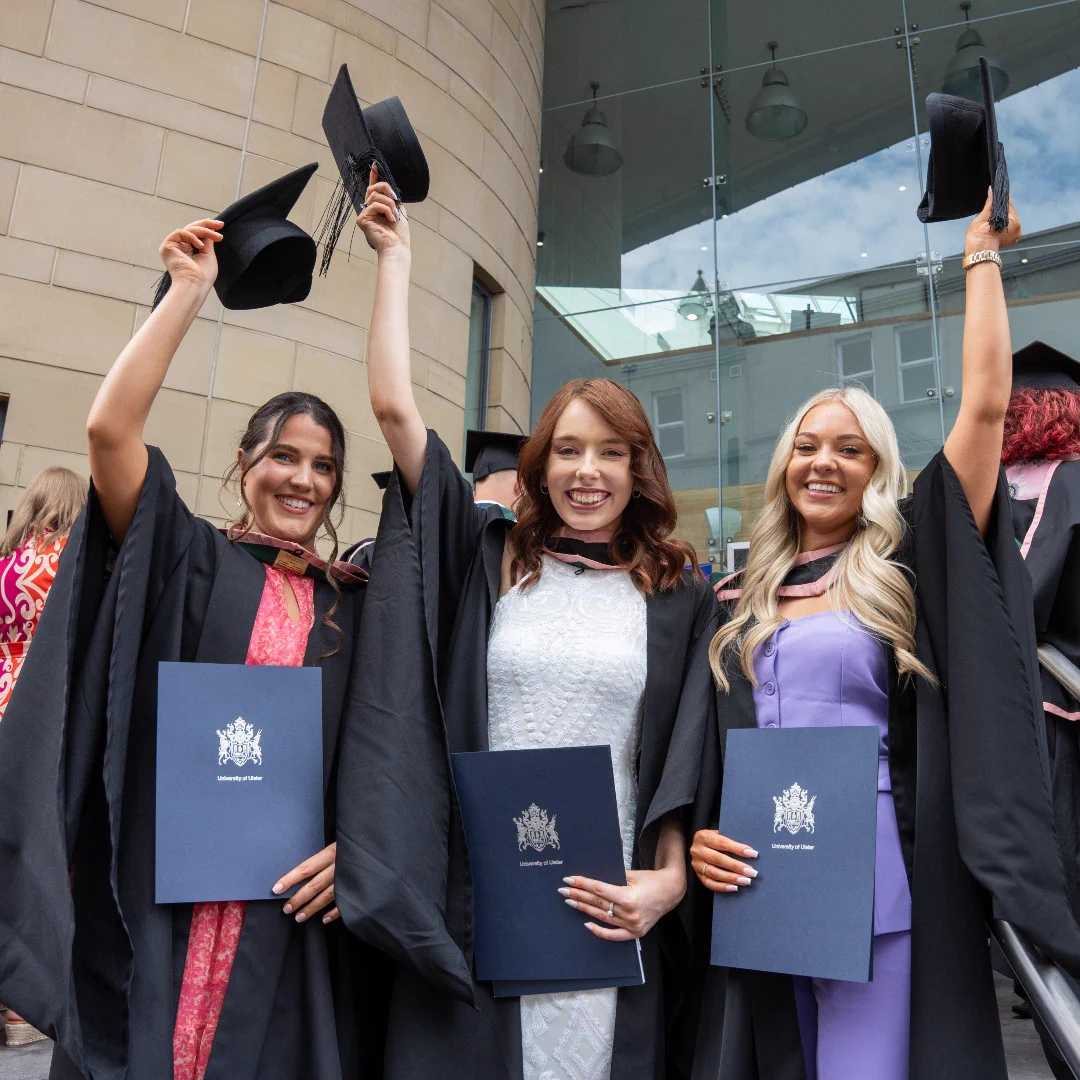 Three graduates in caps and gowns proudly hold diplomas and raise their mortarboards outside a modern university building.